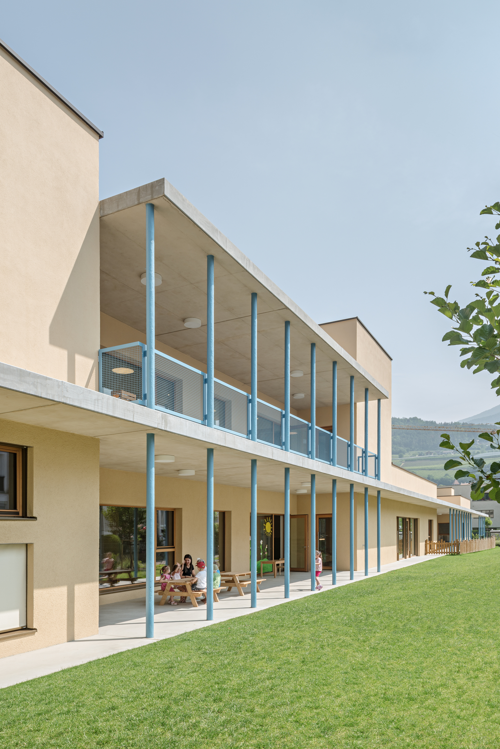 Two-story glass colonnade with slender blue supports facing a sloped lawn with children playing inside