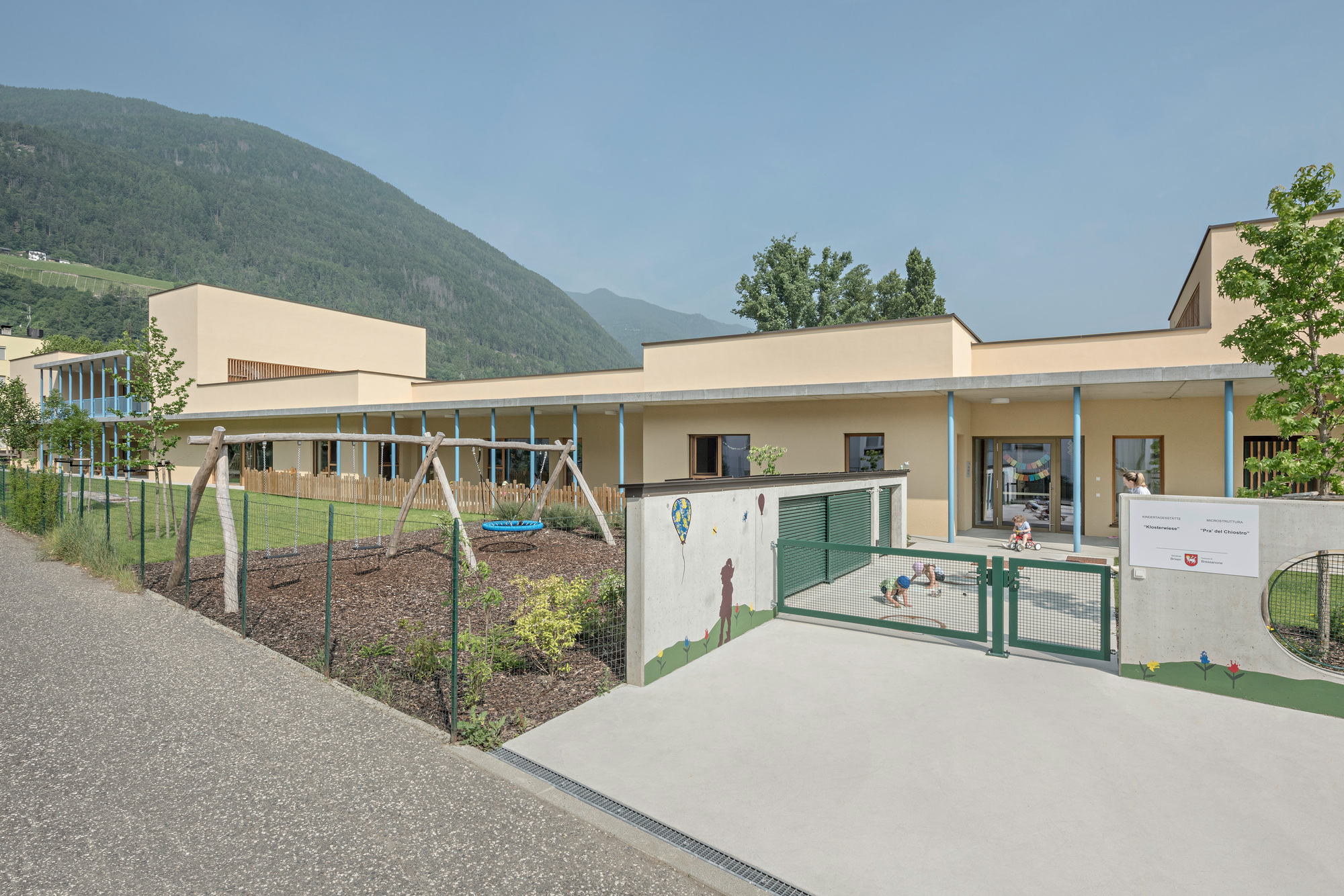 Low-slung building with green entry gates and playground area backed by wooded mountains