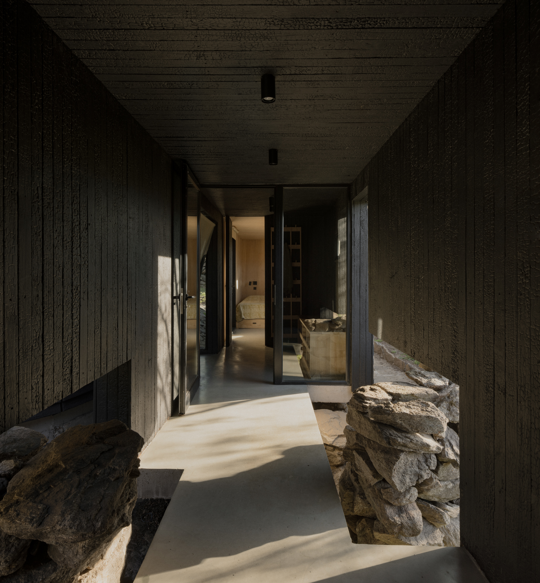 Charred timber corridor with natural light falling across concrete floor and stone walls flanking the passage
