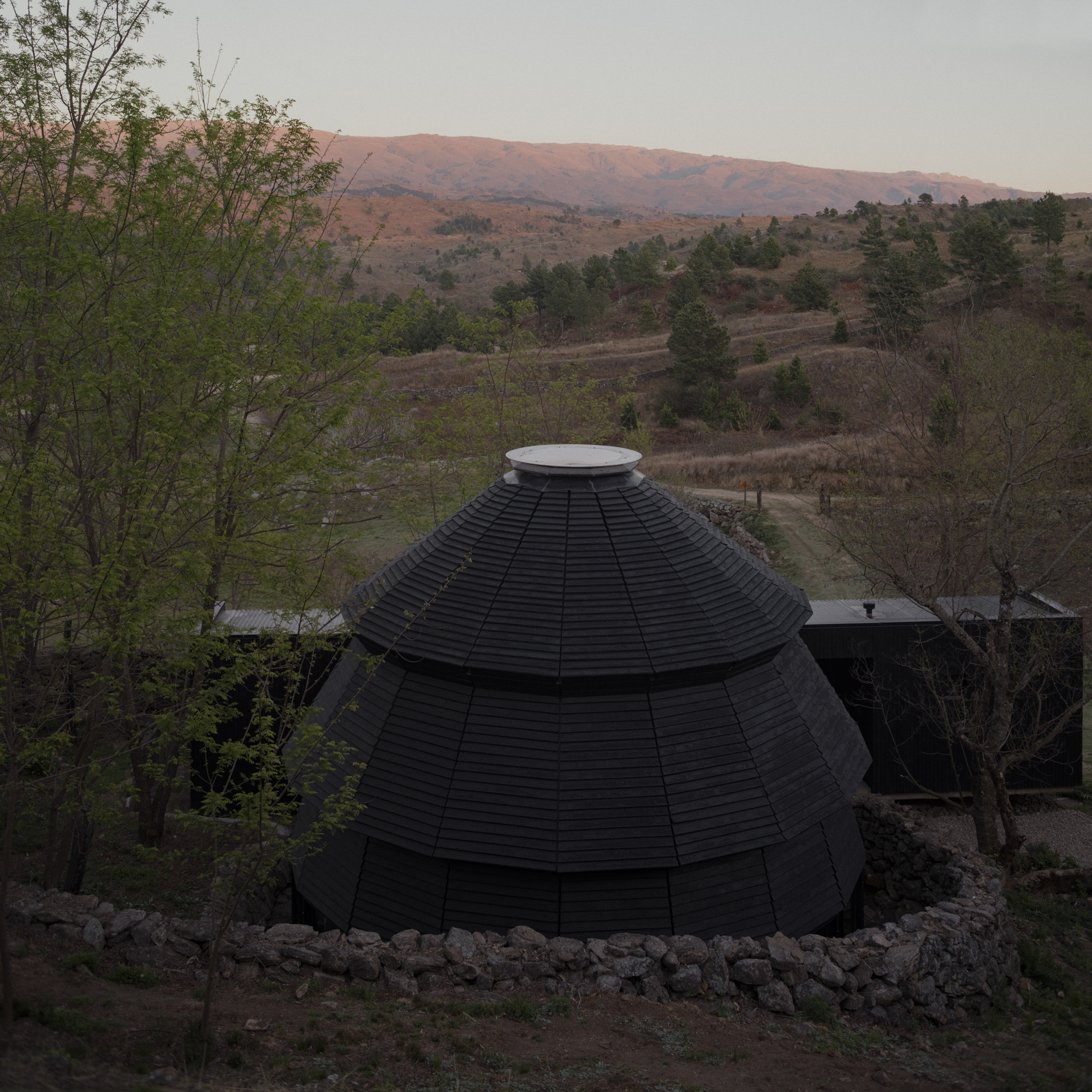 Aerial view of the black shingled dome with circular skylight surrounded by rolling hillside at dusk