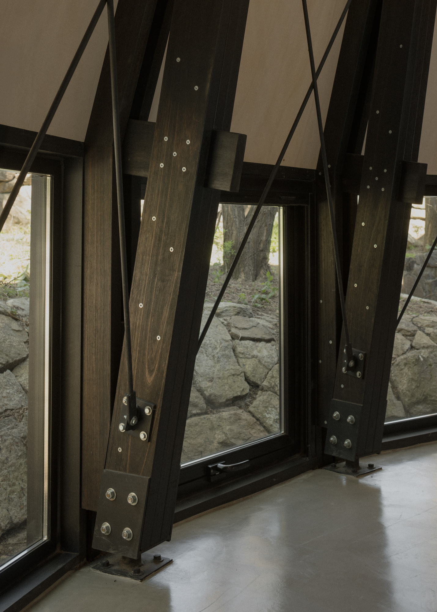 Diagonal timber bracing with bolted steel connections framing a window to a stone wall outside