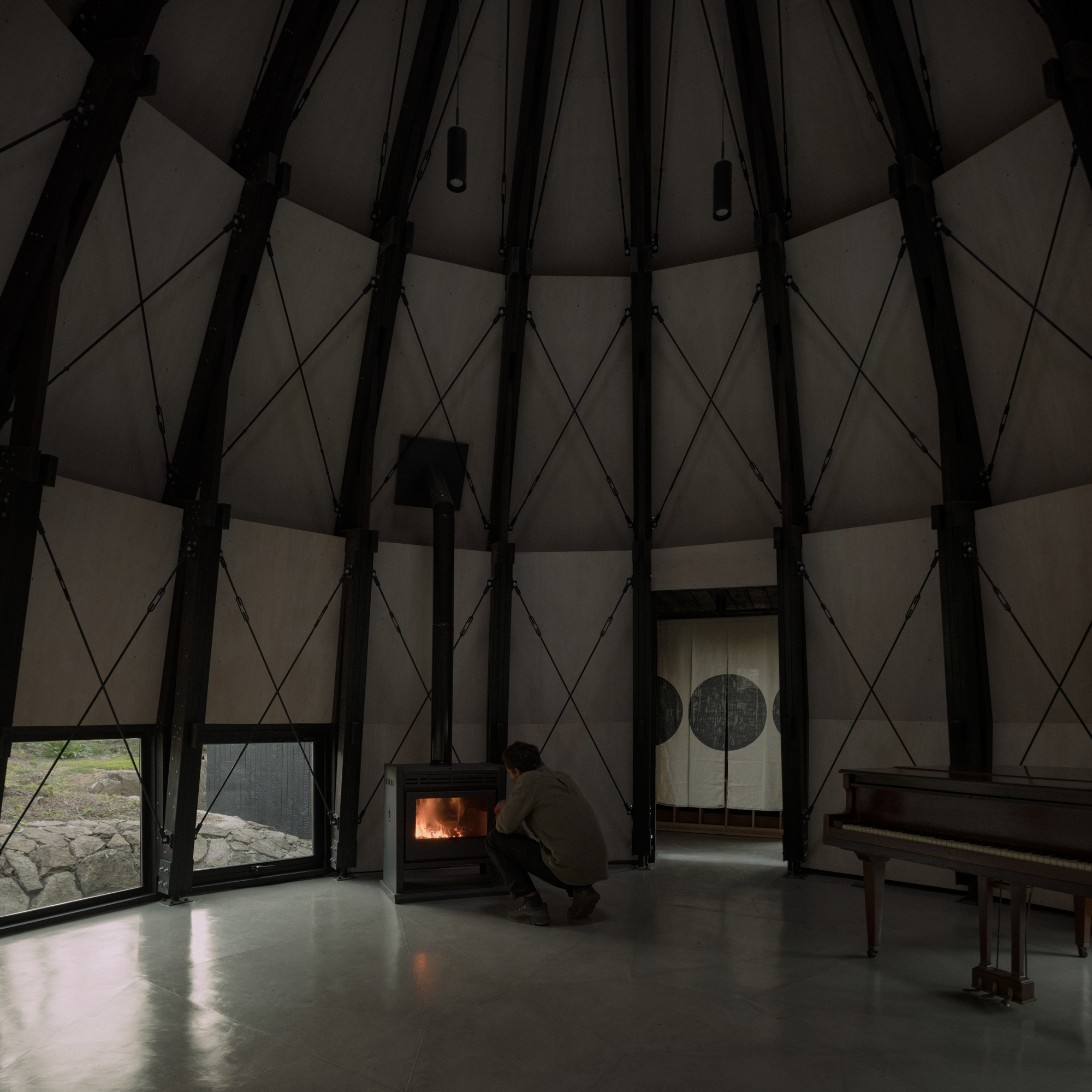 Conical interior with exposed timber ribs as a person crouches beside a glowing wood stove