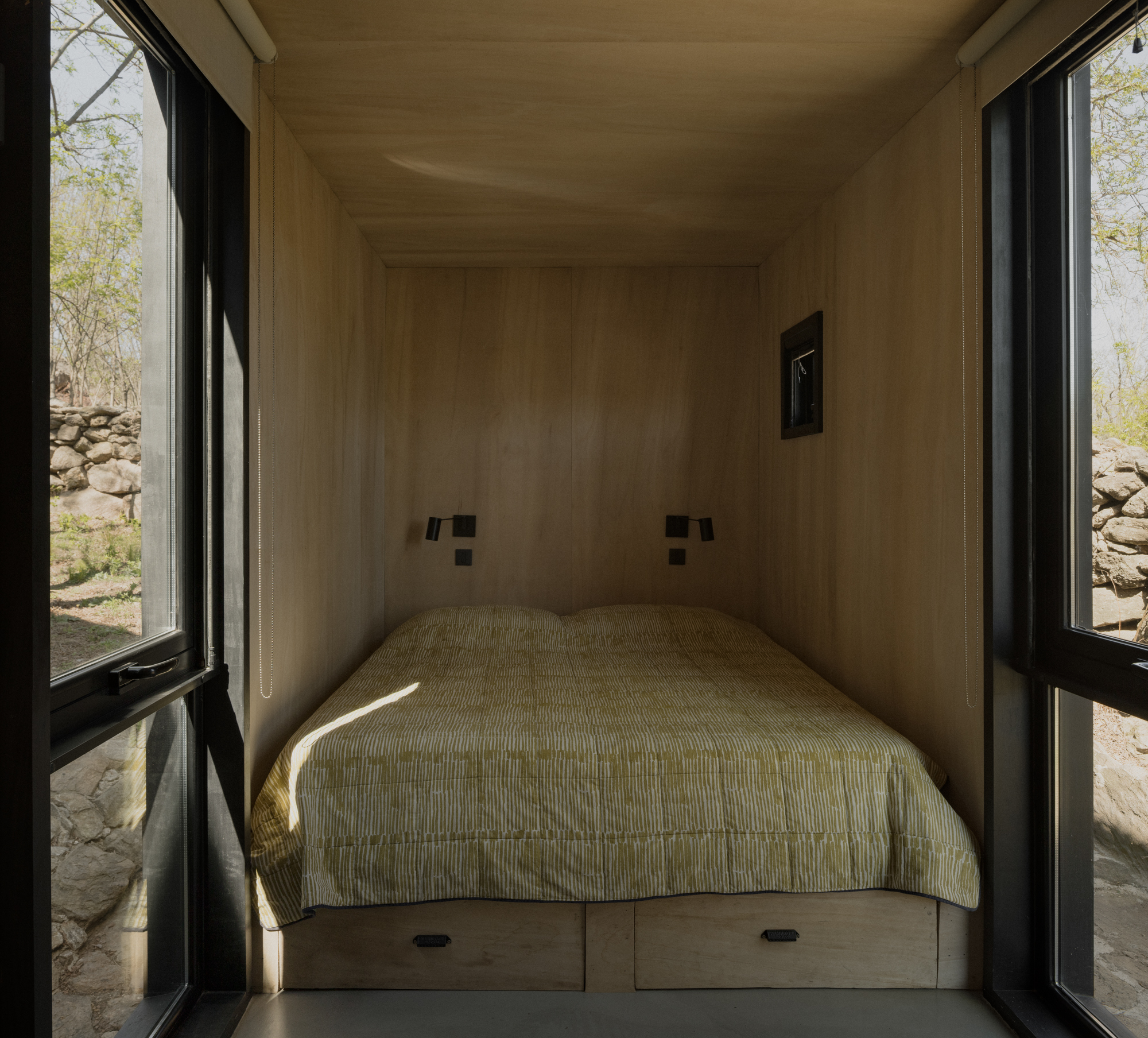 Built-in bed alcove with plywood lining and flanking black-framed glazed doors opening to a stone wall