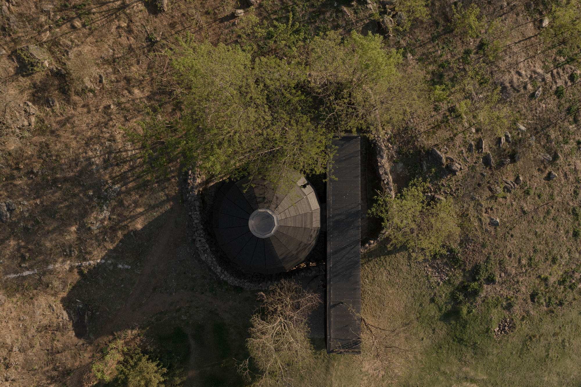 Aerial view of a circular thatched roof structure with stone perimeter wall and linear wing in dry grassland