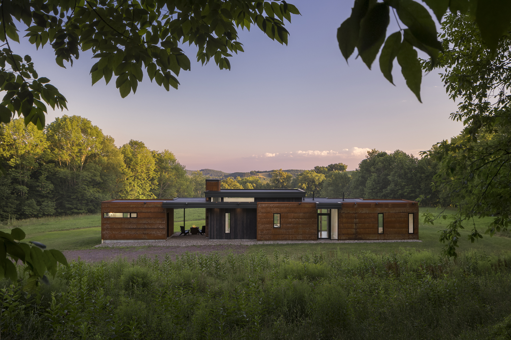 Front facade with horizontal wood cladding and dark glazed sections framed by overhanging tree branches at dusk