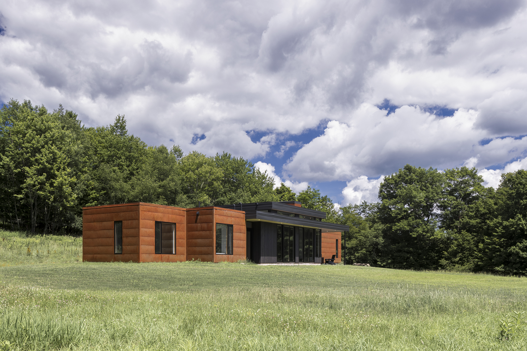 Side elevation showing timber-clad volumes and glazed living area opening to mowed lawn under dramatic clouds