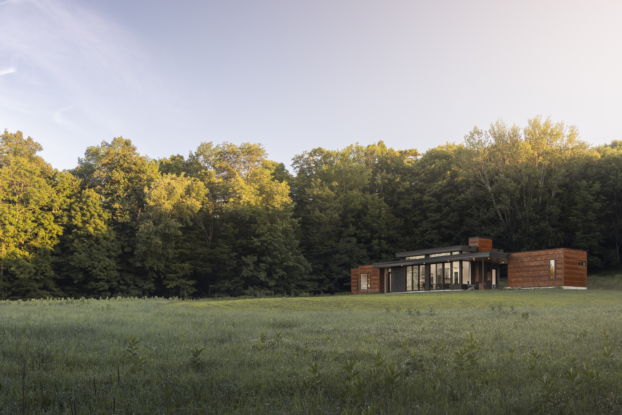 Long view across meadow of the horizontal residence with wood siding and flat roof at sunset