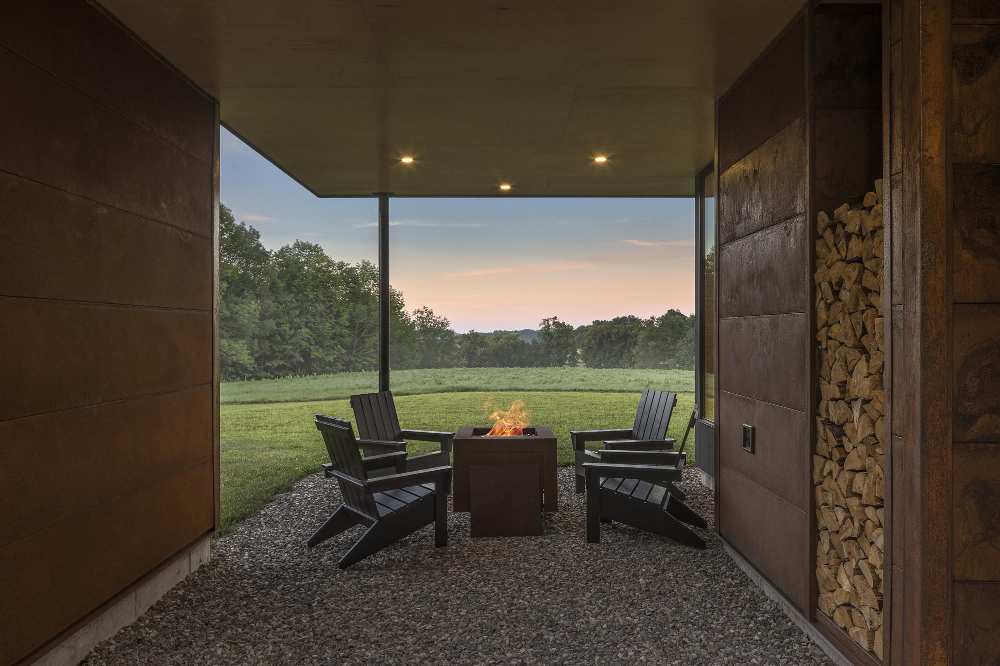 Covered outdoor terrace with fire pit and chairs on gravel looking toward open field at twilight