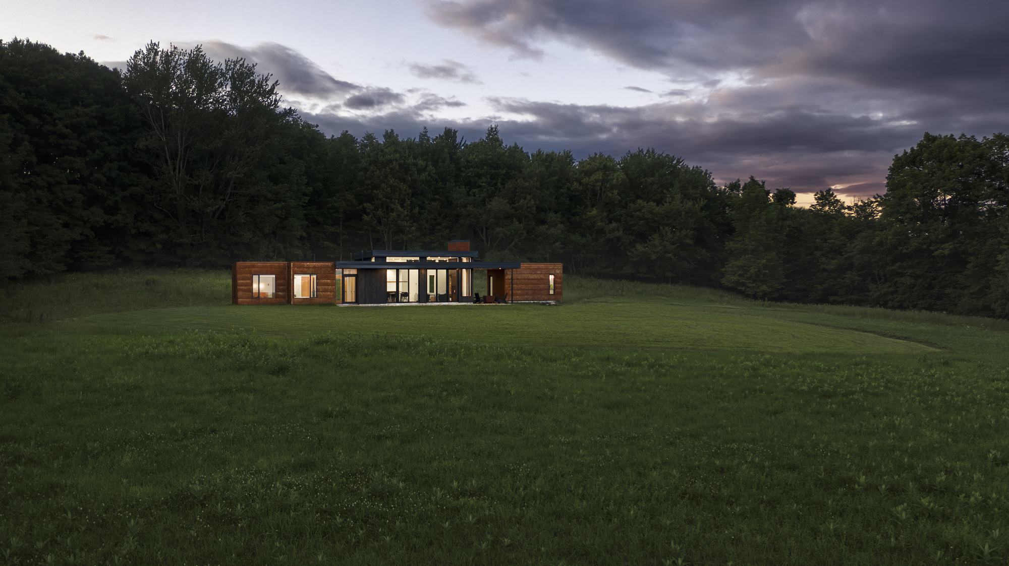 Distant view of the horizontal timber and glass volume set in a meadow at dusk