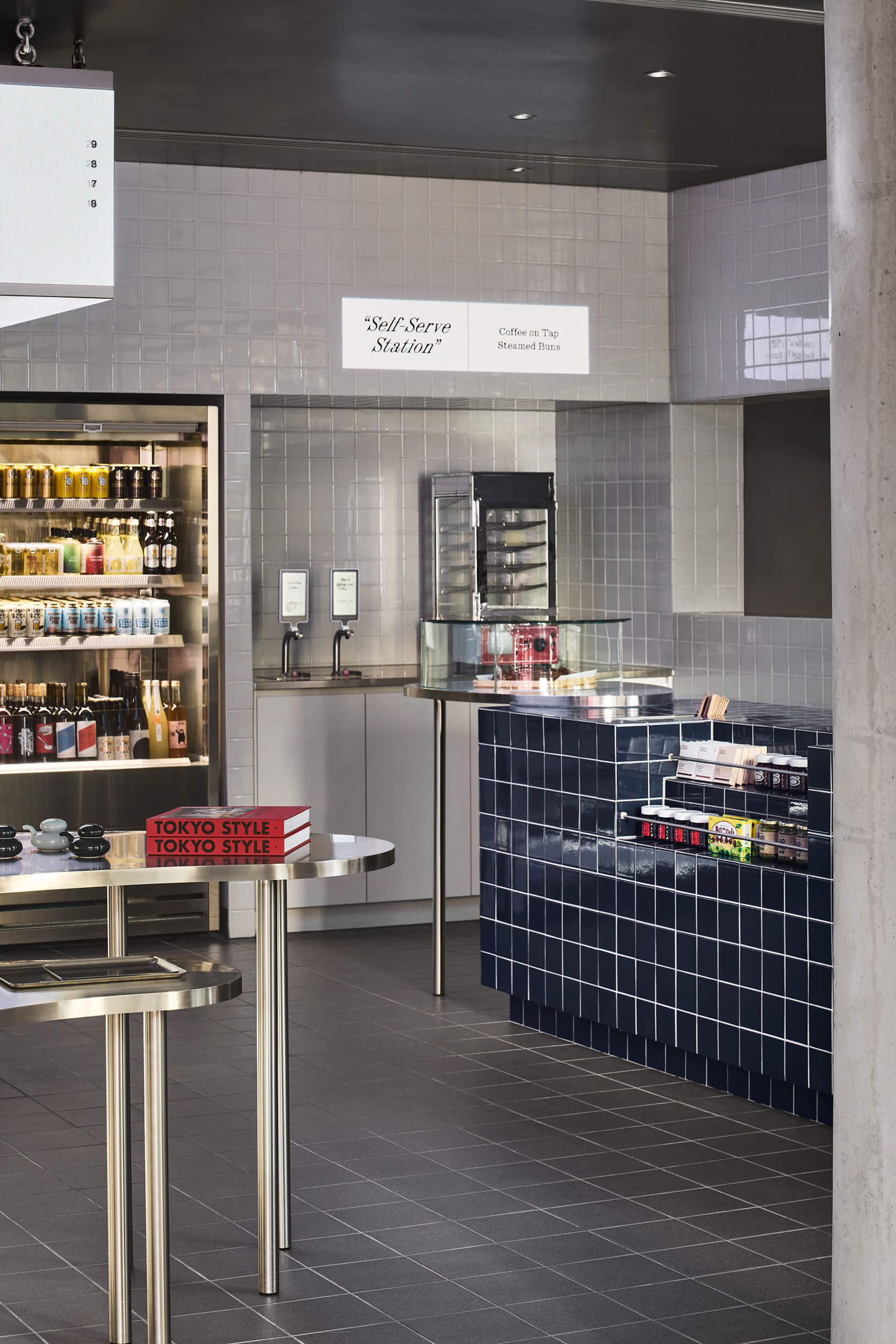 Self-serve station with navy tiled counter and illuminated shelving visible beyond glass vitrine and concrete column