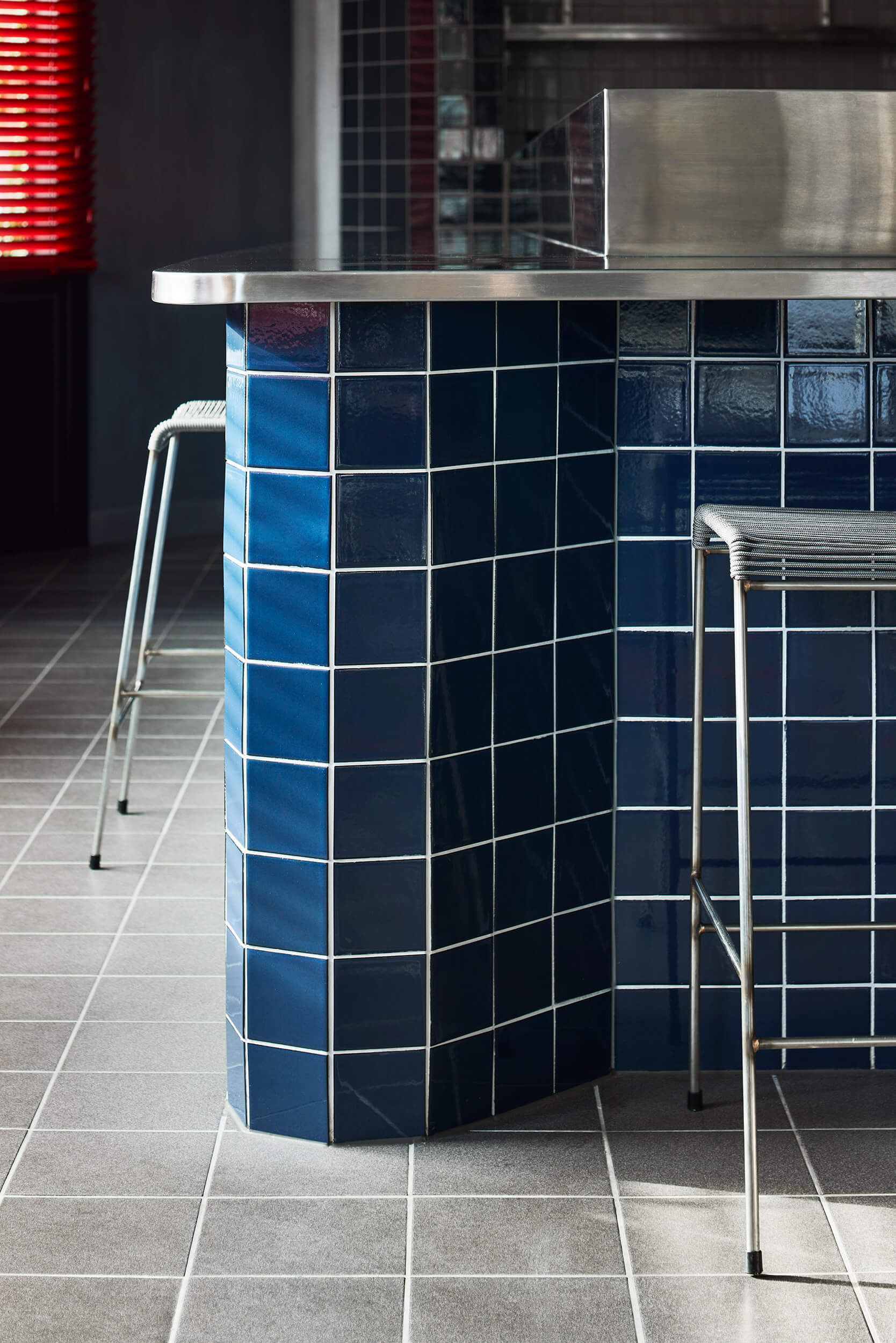 Curved blue tiled counter edge with stainless steel top and grey floor tile below a stool