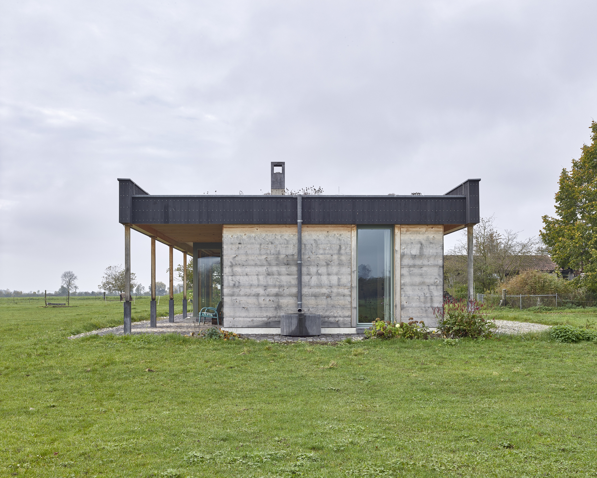Board-formed concrete pavilion with metal-clad overhanging roof and central chimney in open meadow under overcast sky