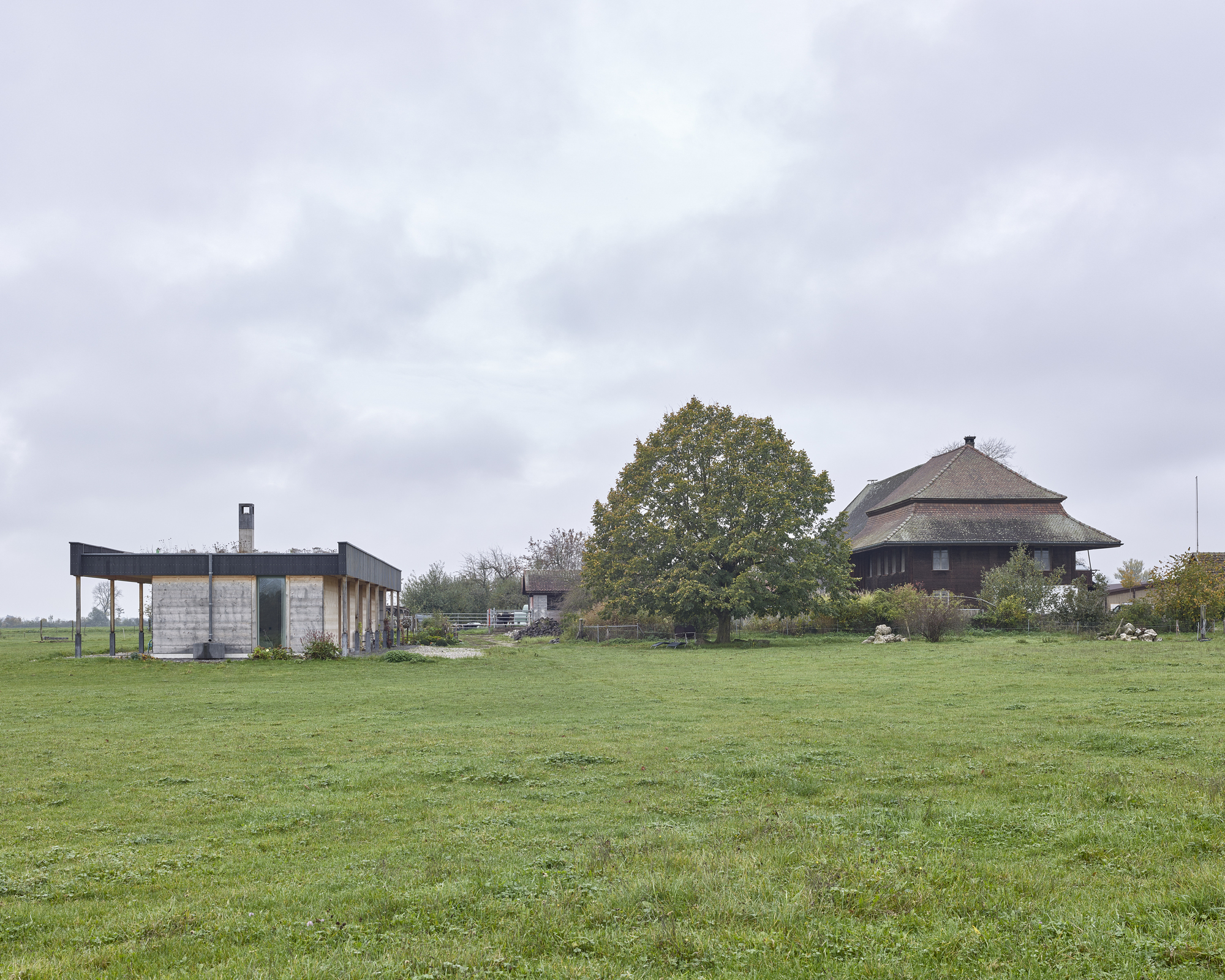 Concrete and steel pavilion beside traditional thatched-roof barn in grassy field under cloudy autumn sky
