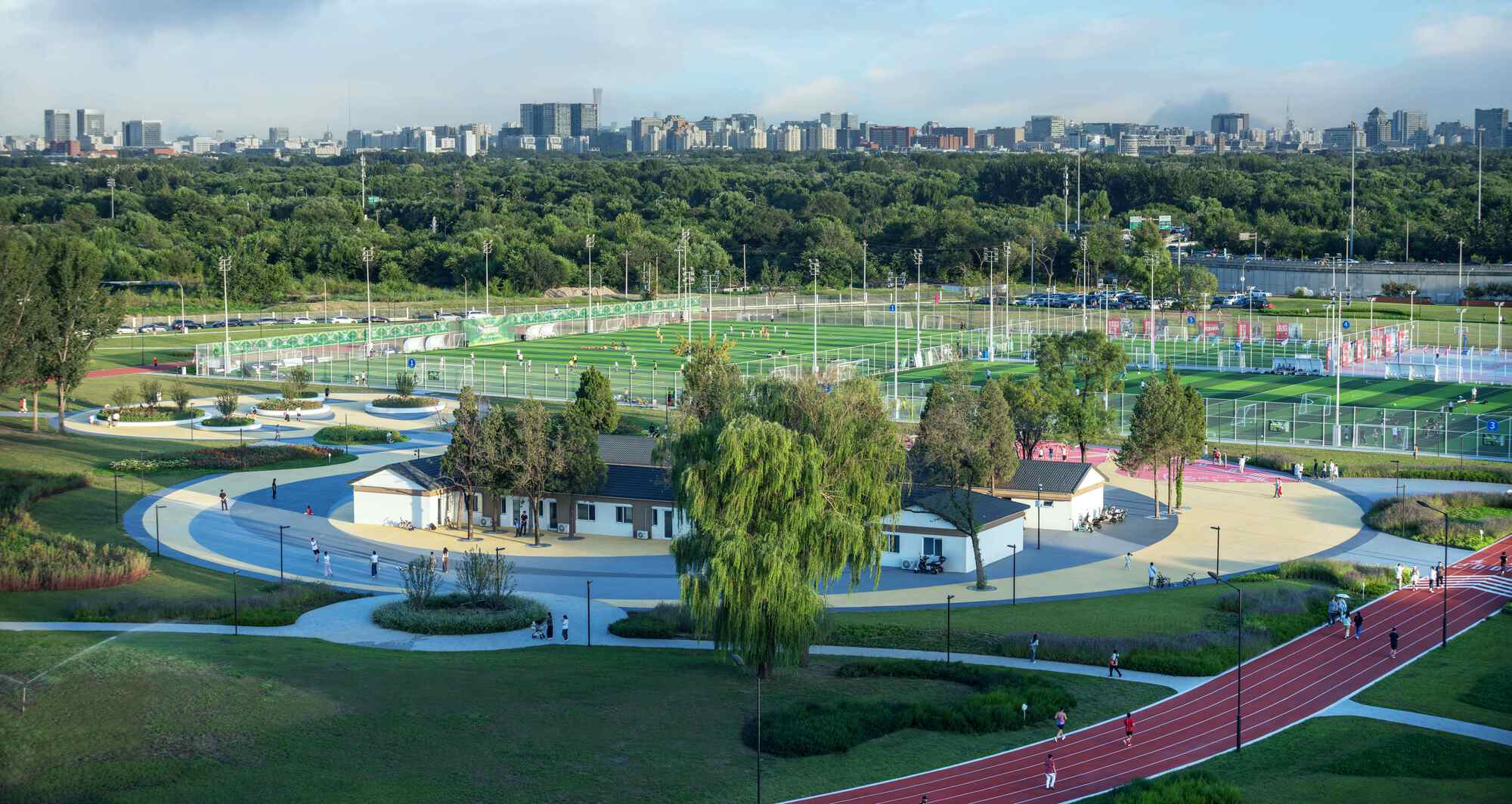 Elevated view of circular pathway surrounding white pavilion with willow trees and sports fields beyond