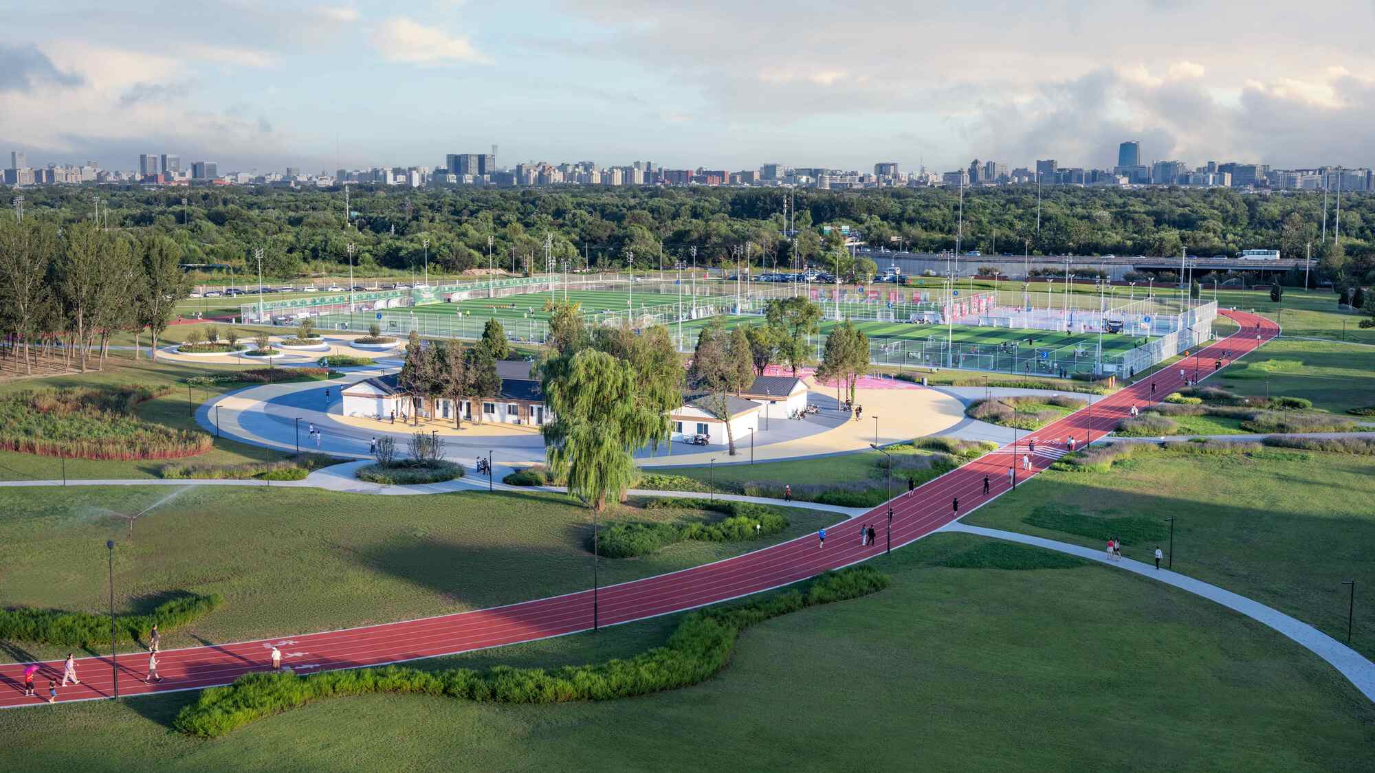 Winding red track alongside curved white path through parkland with distant city skyline under cloudy sky