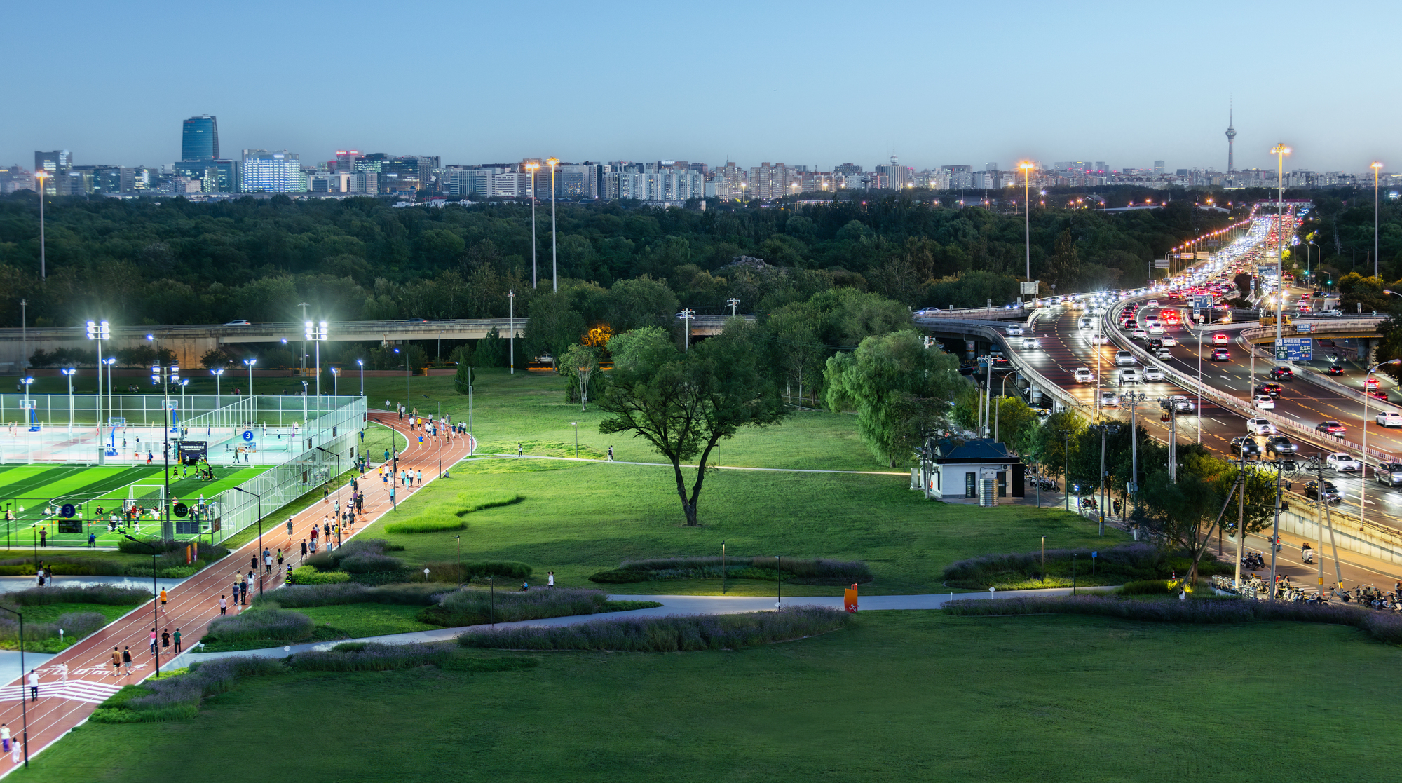 Elevated view of the park at dusk with running track, sports courts, and highway traffic beyond