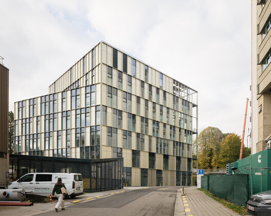 Rear street view: beige and black facade of the centre rising above a parked van, delivery forecourt, autumn trees and adjacent campus block at right
