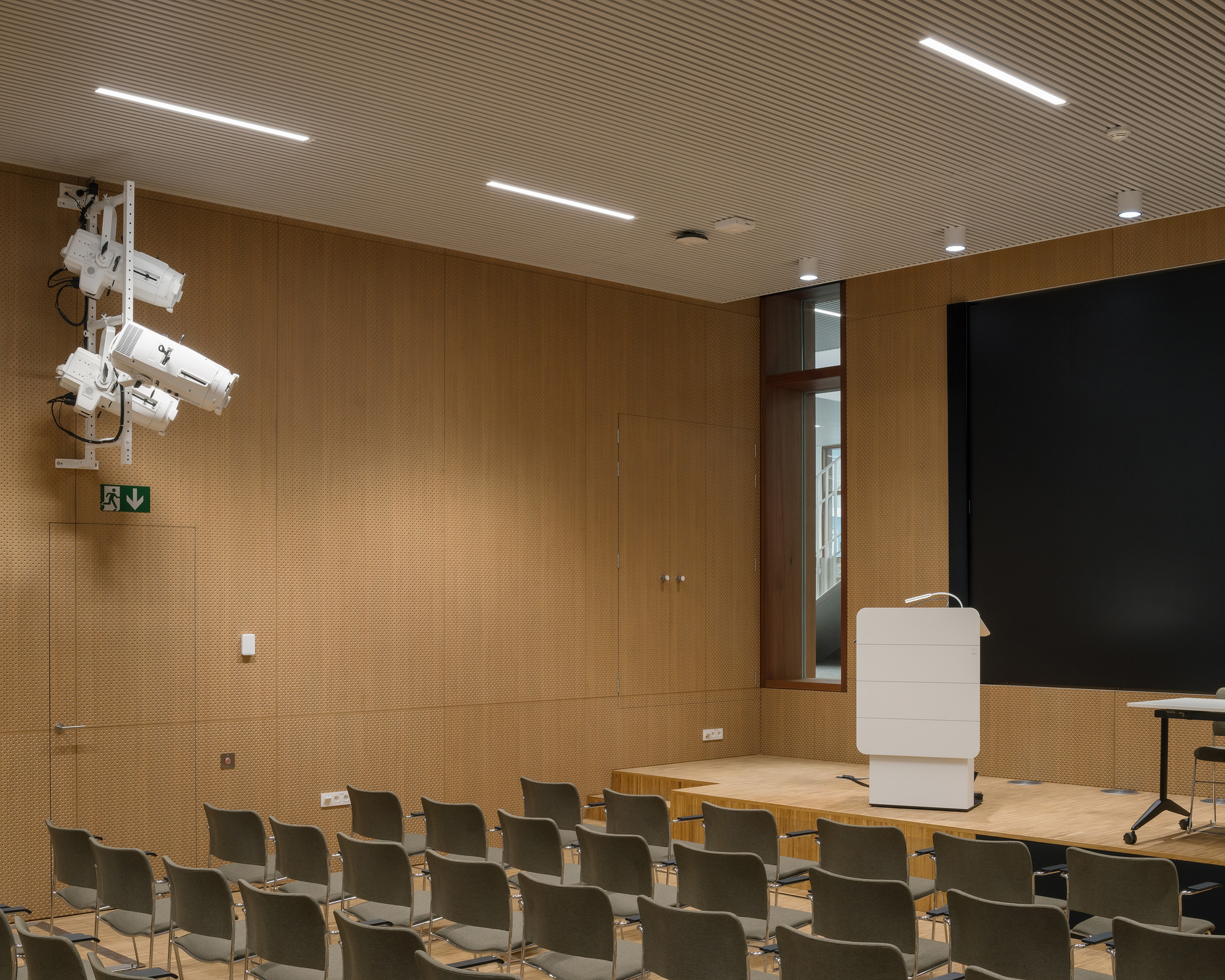 Auditorium: timber-clad walls and ceiling, black projection screen, illuminated white lectern on a low timber stage, recessed linear lights, rows of dark chairs