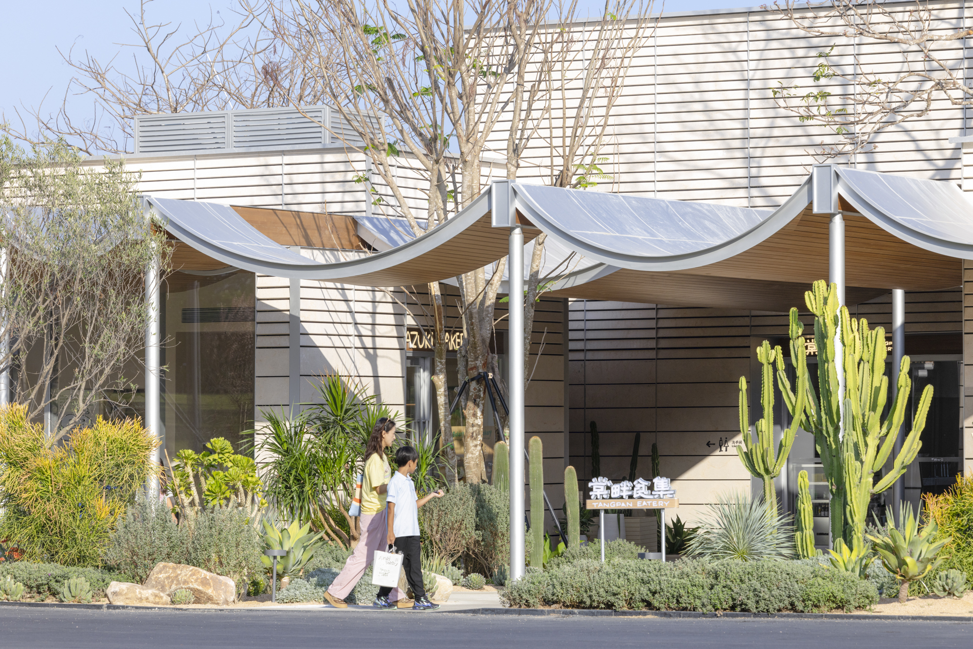 Two visitors walking beneath a scalloped fabric canopy beside a horizontal wood clad facade