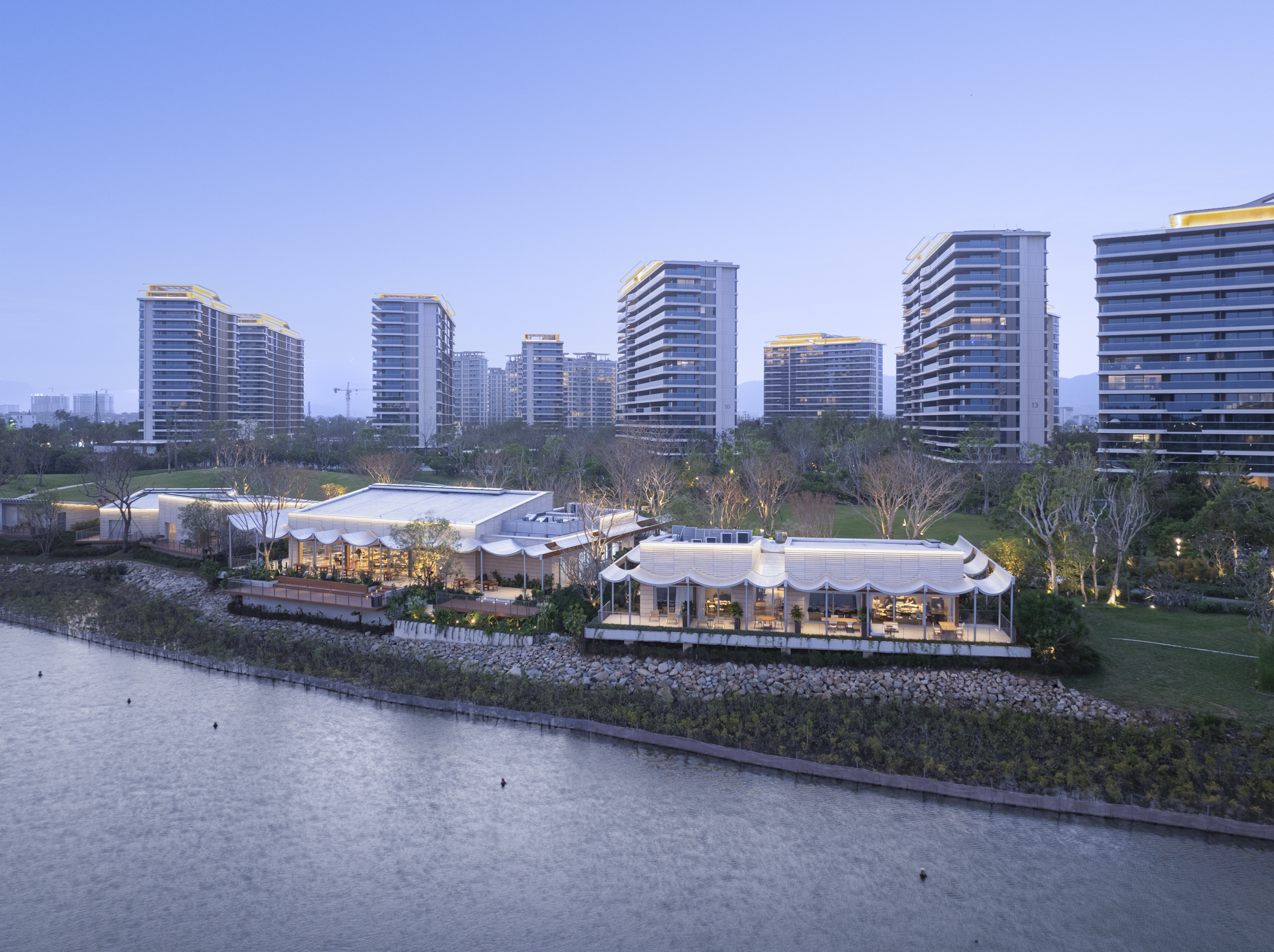 Evening view of white undulating roofs along the lake edge with illuminated high-rise towers beyond