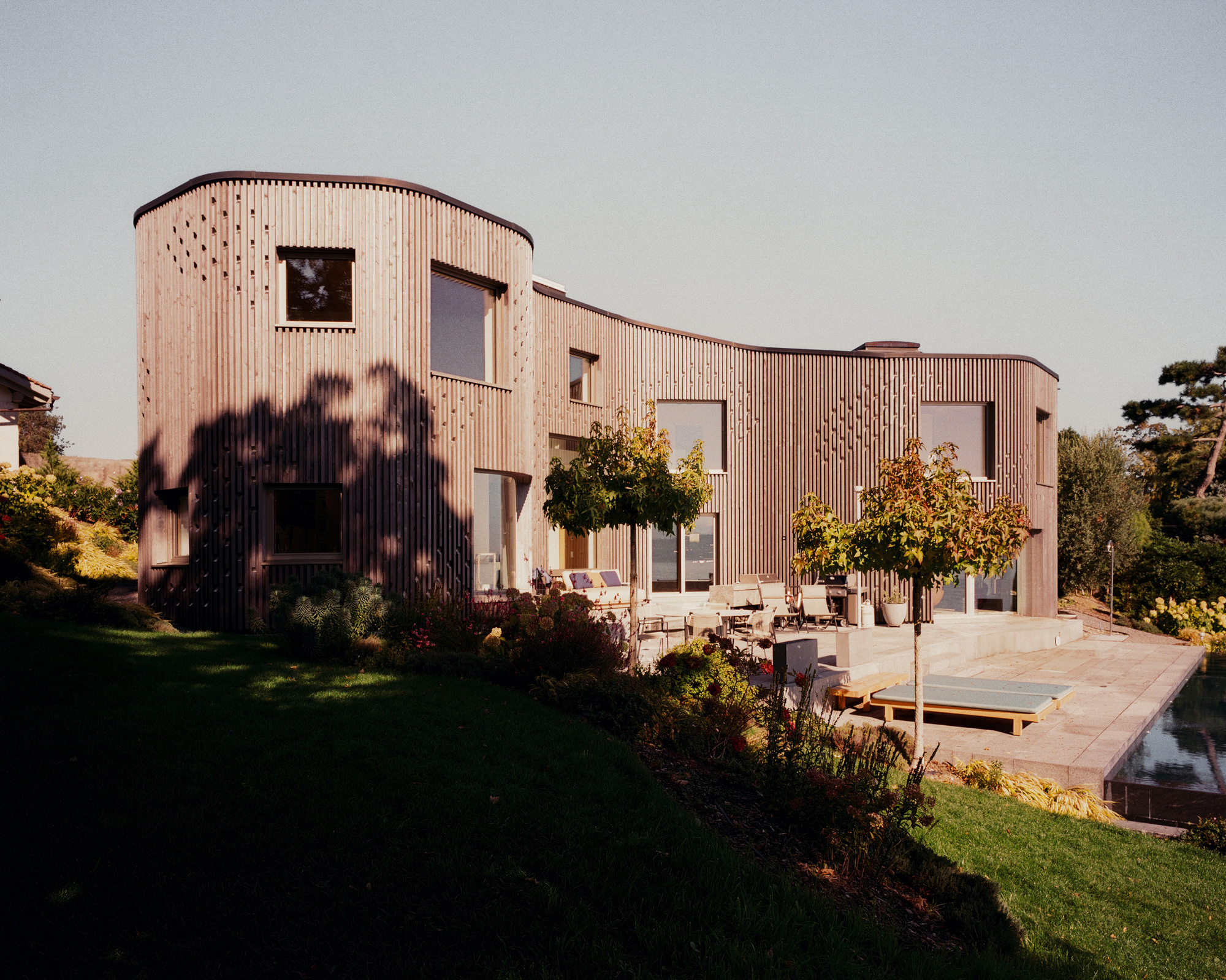 Curved timber-clad facade with carved openings overlooking a garden terrace at sunset