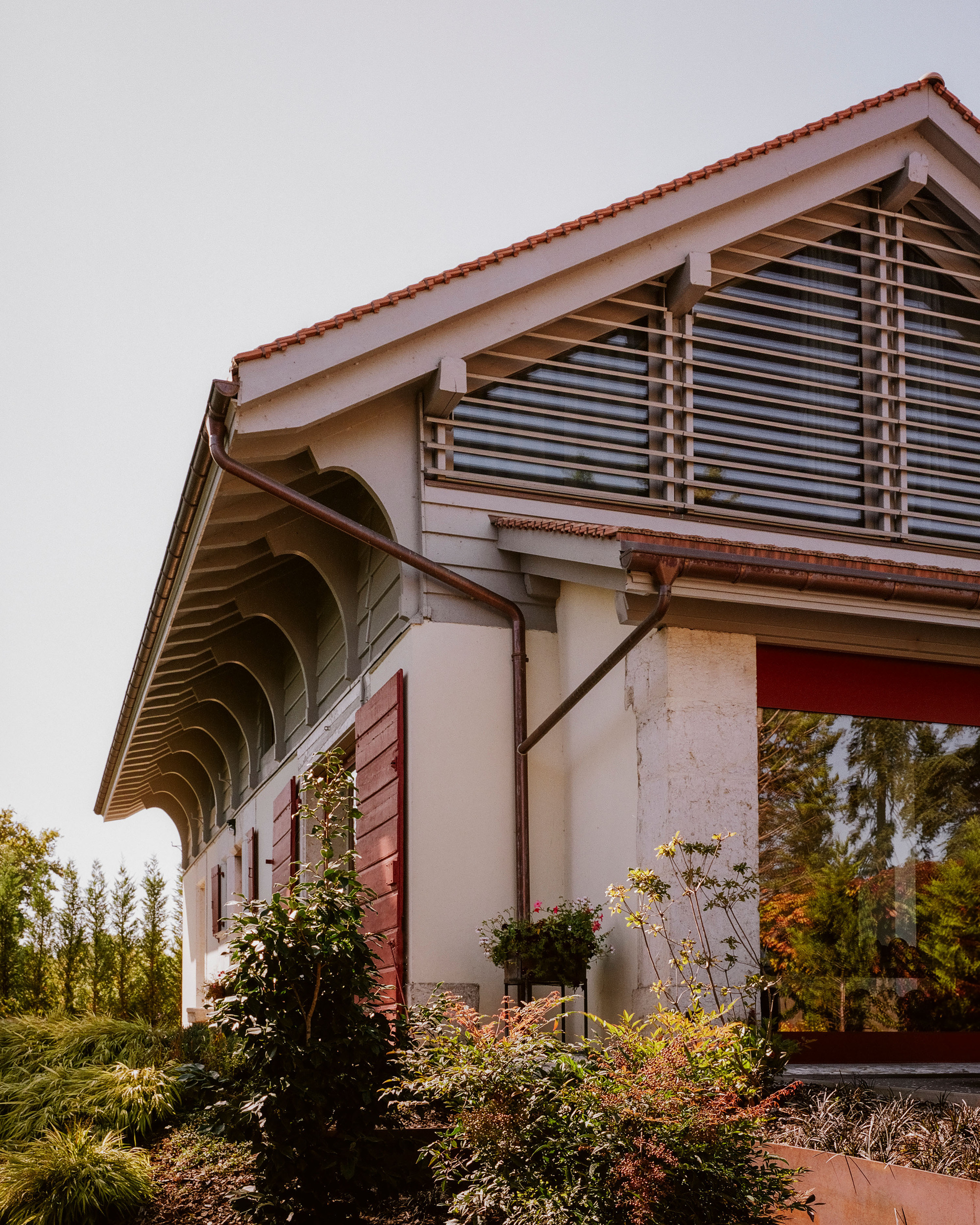 Gabled facade with red tile roof, horizontal louvered shutters, and planted garden bed below