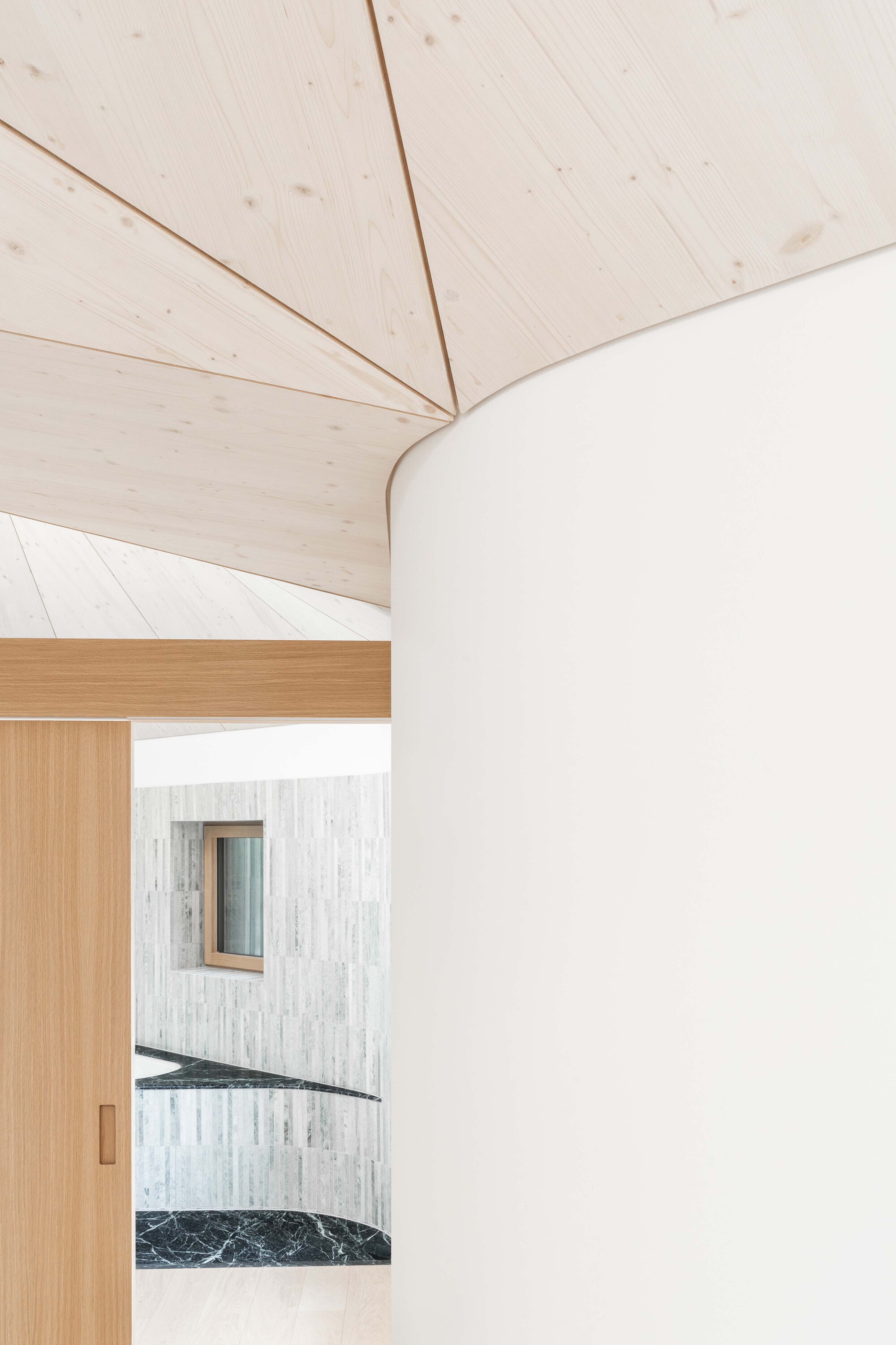 Detail of pale timber planked vaulted ceiling meeting white curved wall above timber doorframe