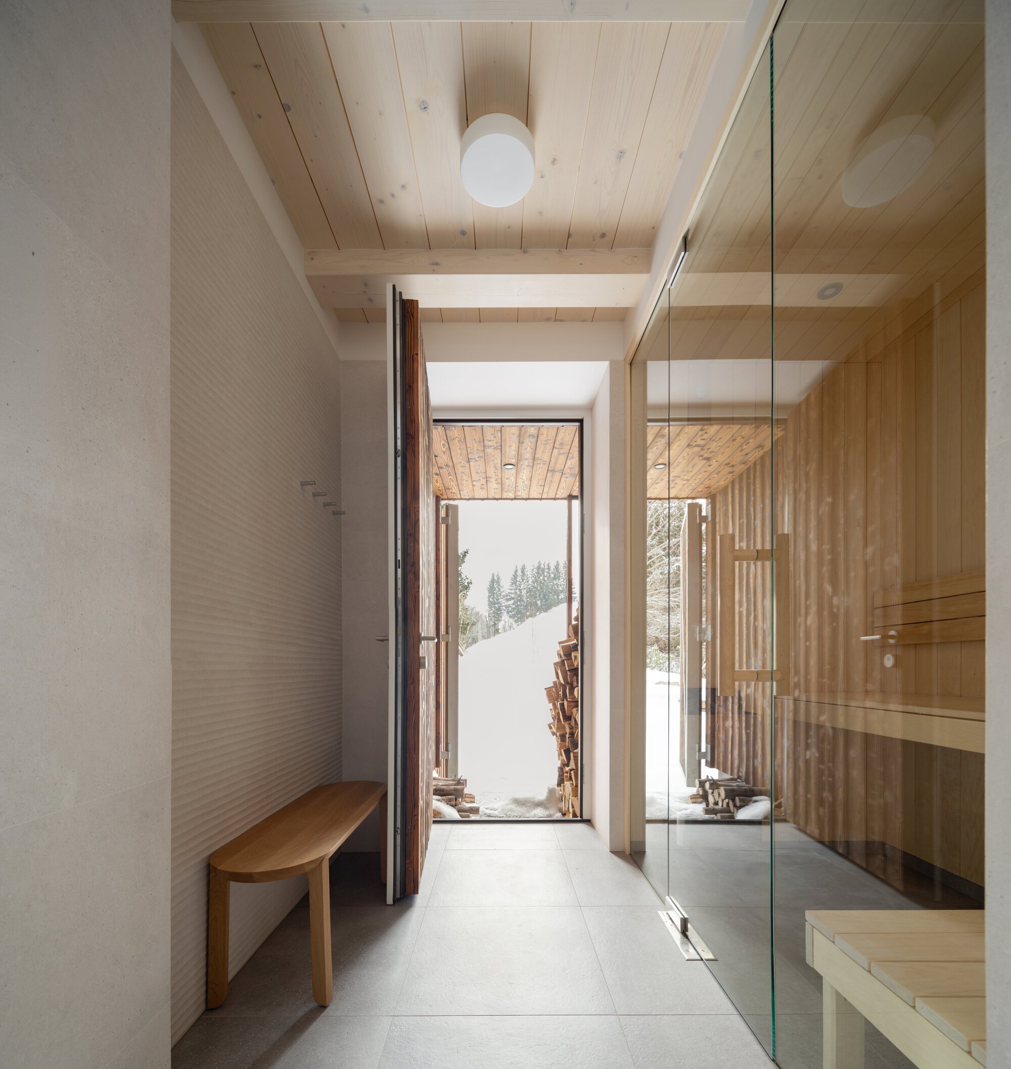 Timber-lined entryway with glass sauna enclosure and open door framing stacked firewood and snowy landscape beyond
