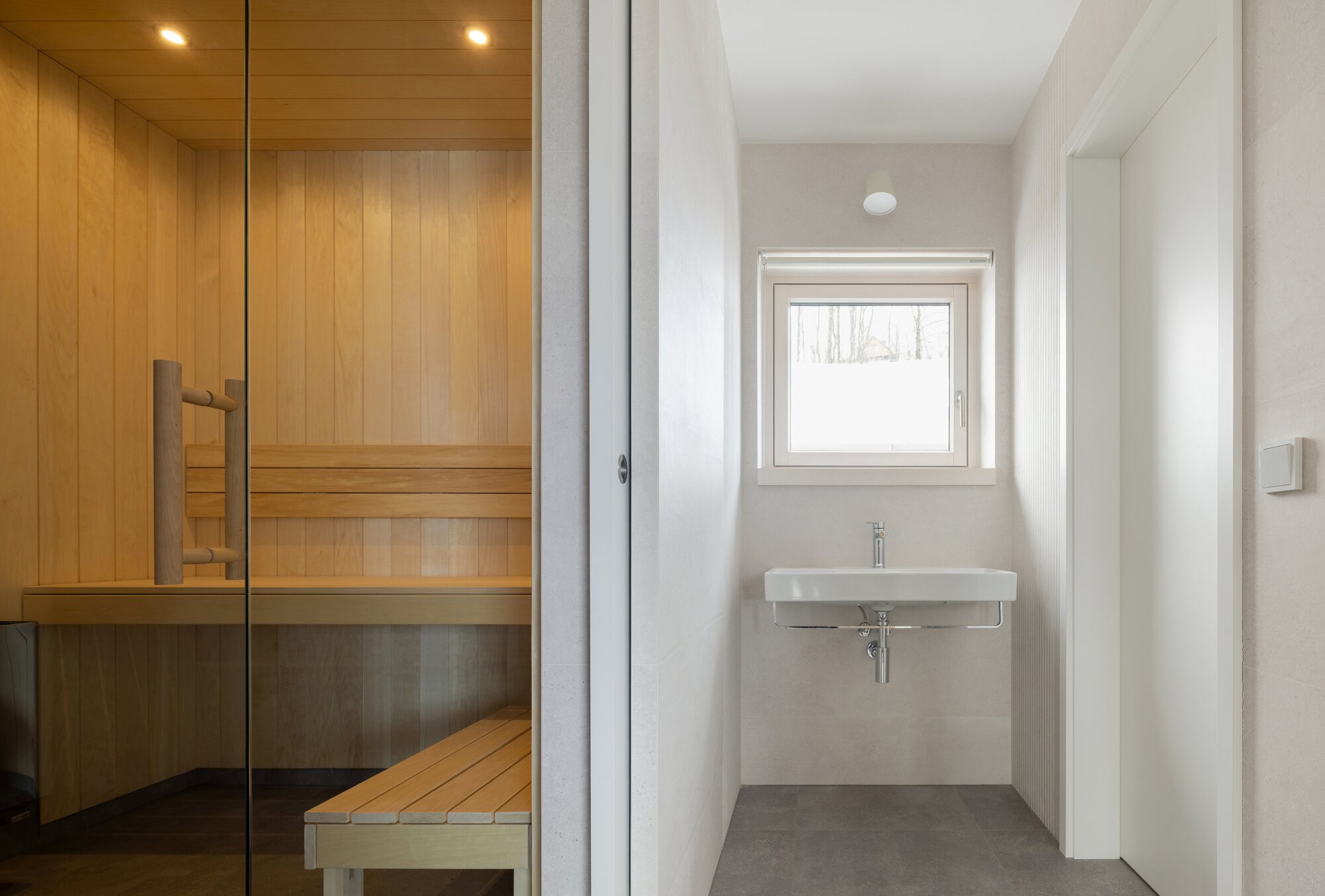 Glass-enclosed sauna with timber benches beside a minimal bathroom with wall-mounted sink and square window