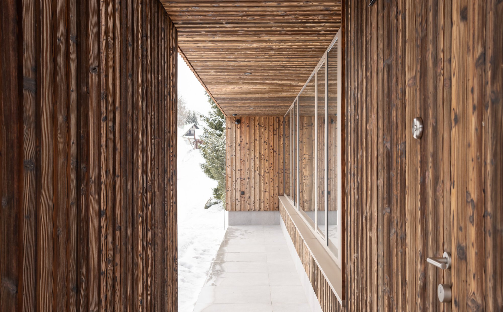 Covered walkway with vertical timber walls, slatted ceiling, and continuous glazing overlooking snowy ground