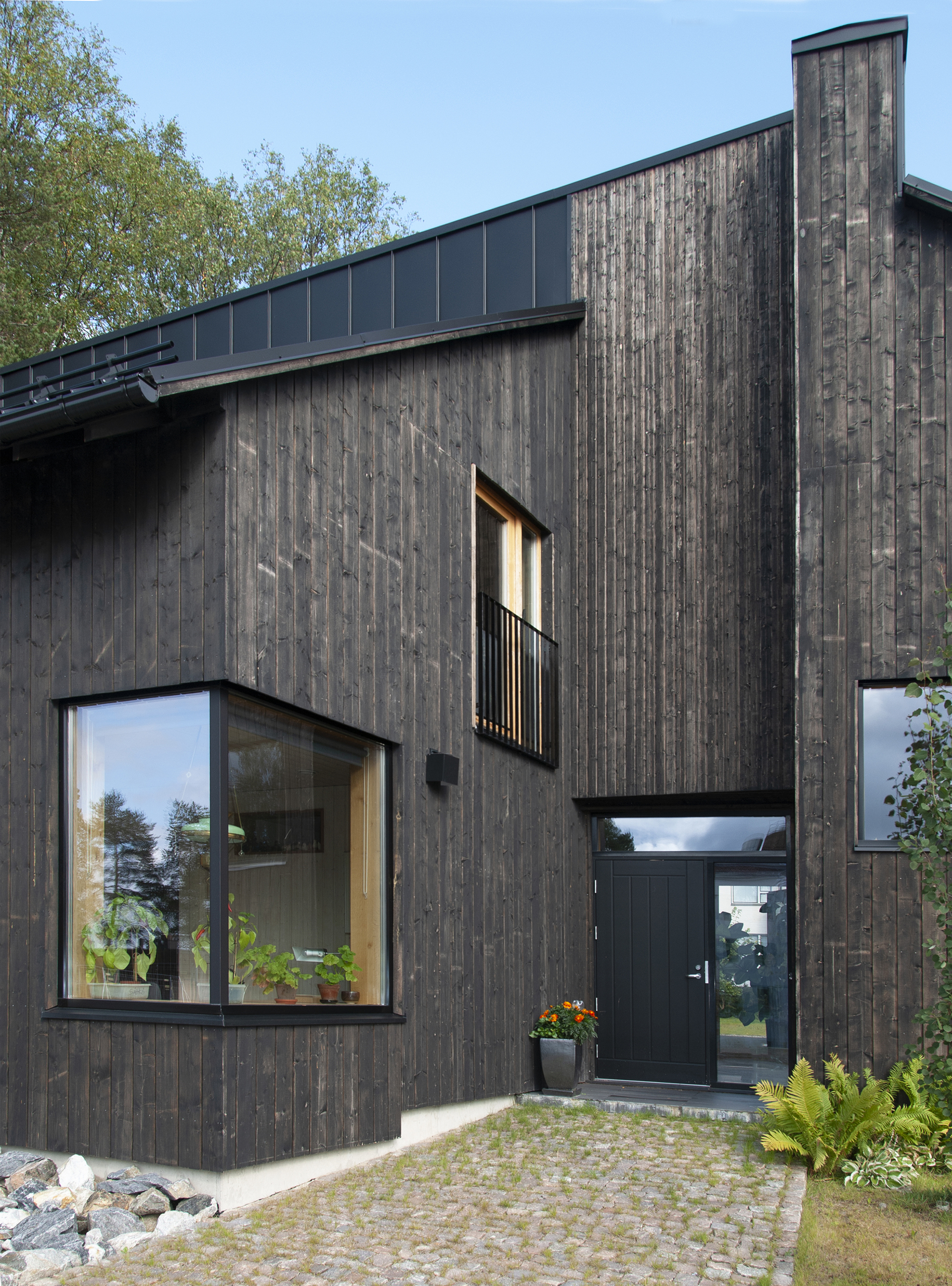 Entrance facade featuring vertical timber boards, projecting window bay with steel railing, and cobblestone paving