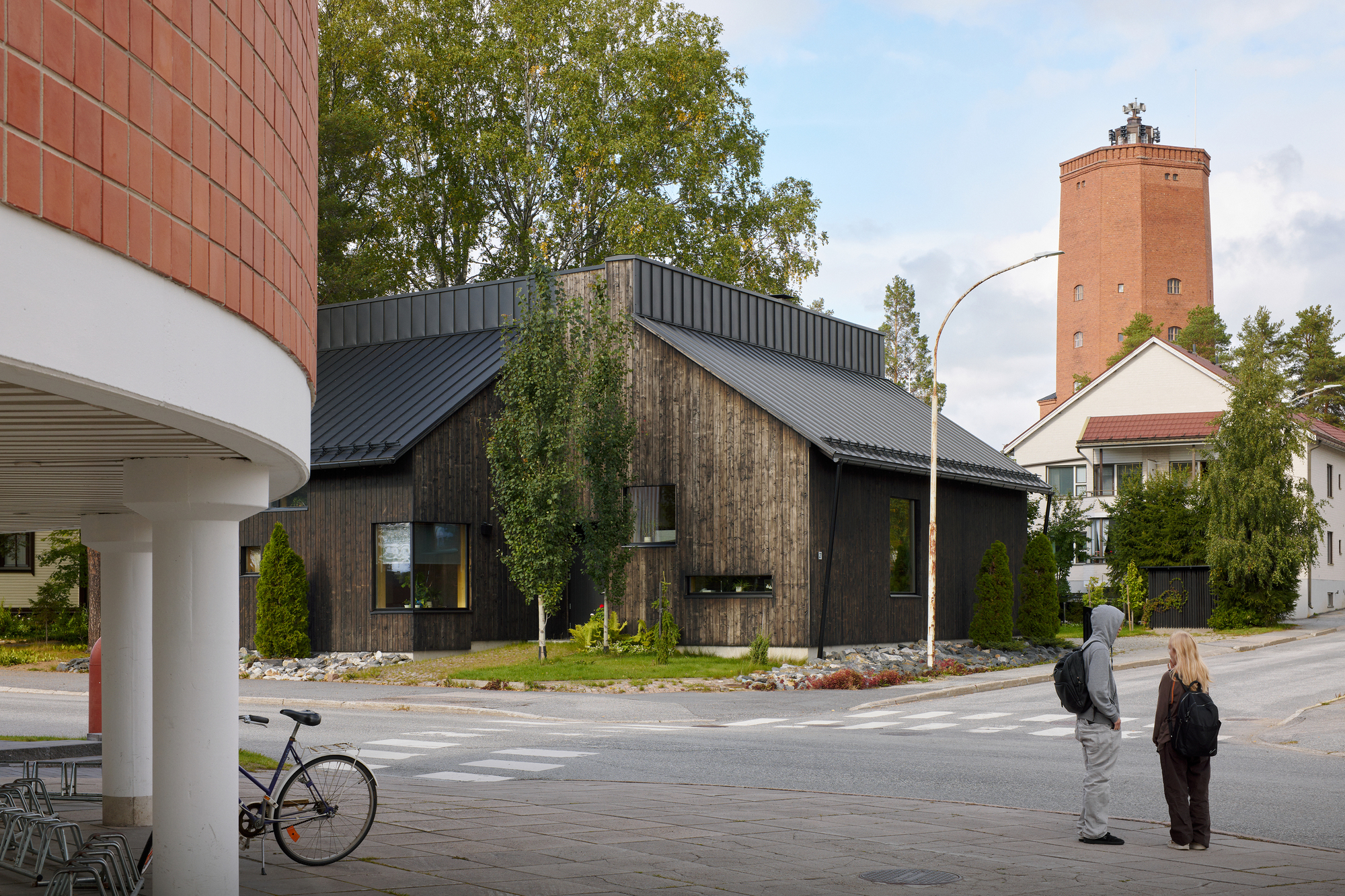 Corner lot view showing timber house with metal roof, water tower, and two pedestrians crossing