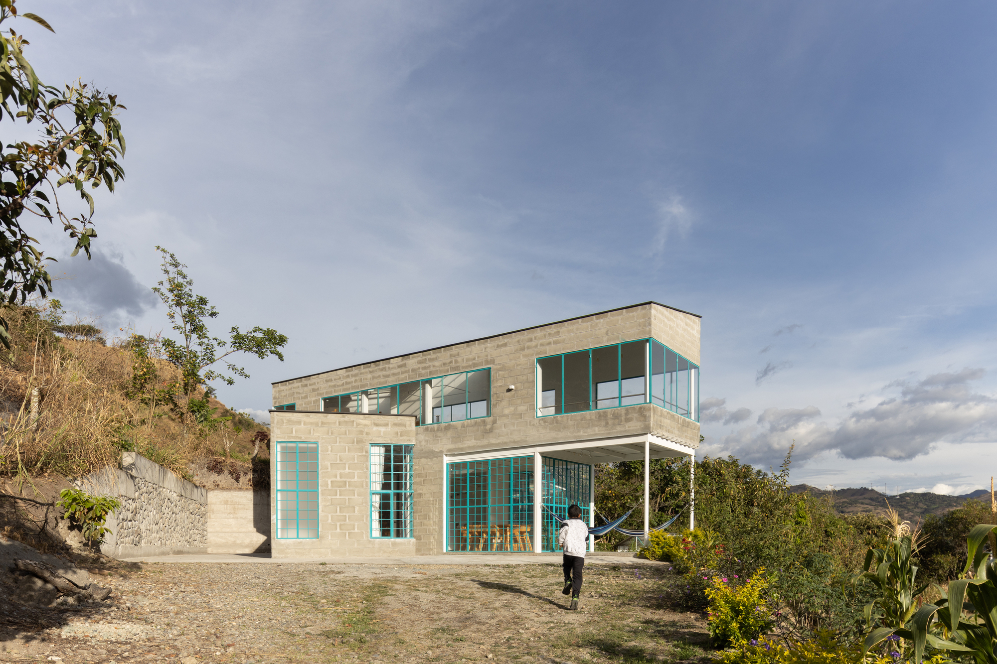 Two-story concrete block house with turquoise-framed windows and a covered terrace on a hillside clearing