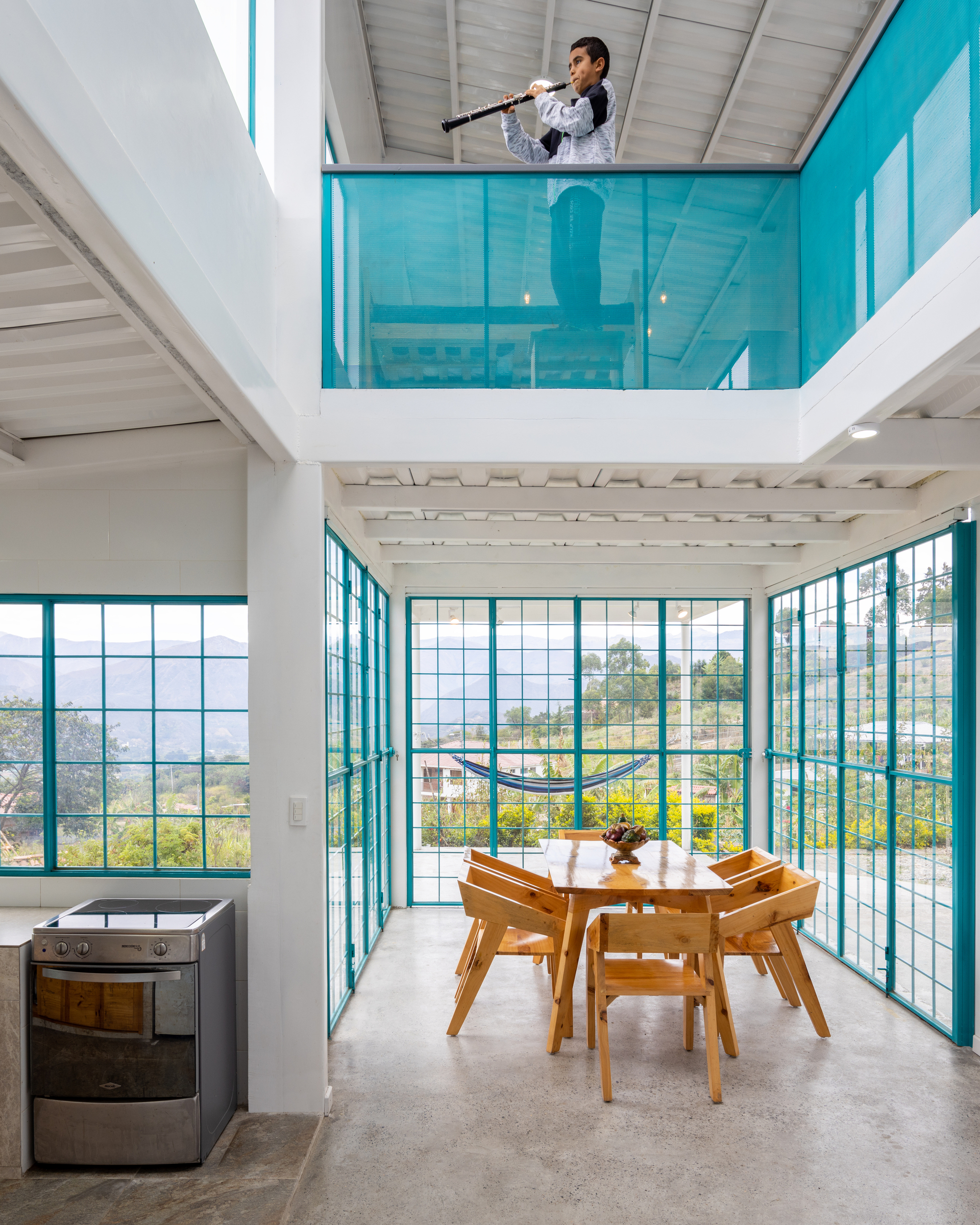 Double-height dining area with turquoise-framed glazing and a mezzanine balcony above timber furniture