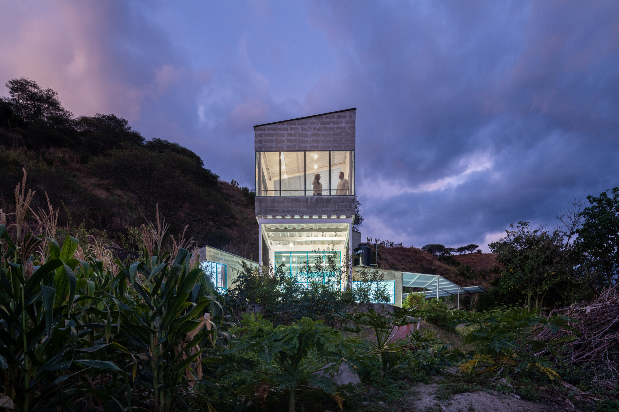 Illuminated tower volume at dusk with a figure visible through the transparent upper level
