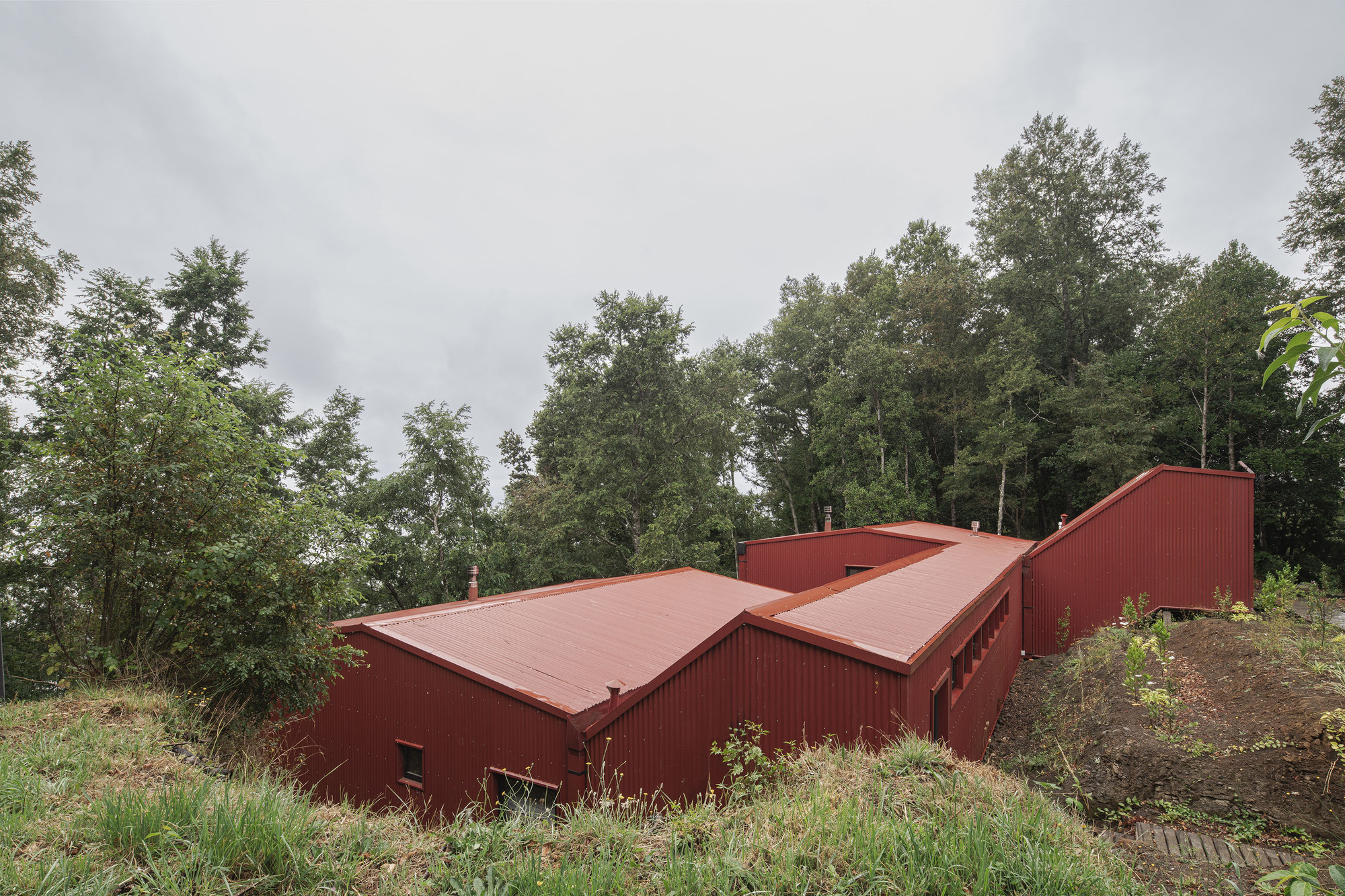 Aerial view of clustered red metal volumes with gabled roofs nestled into the forested slope