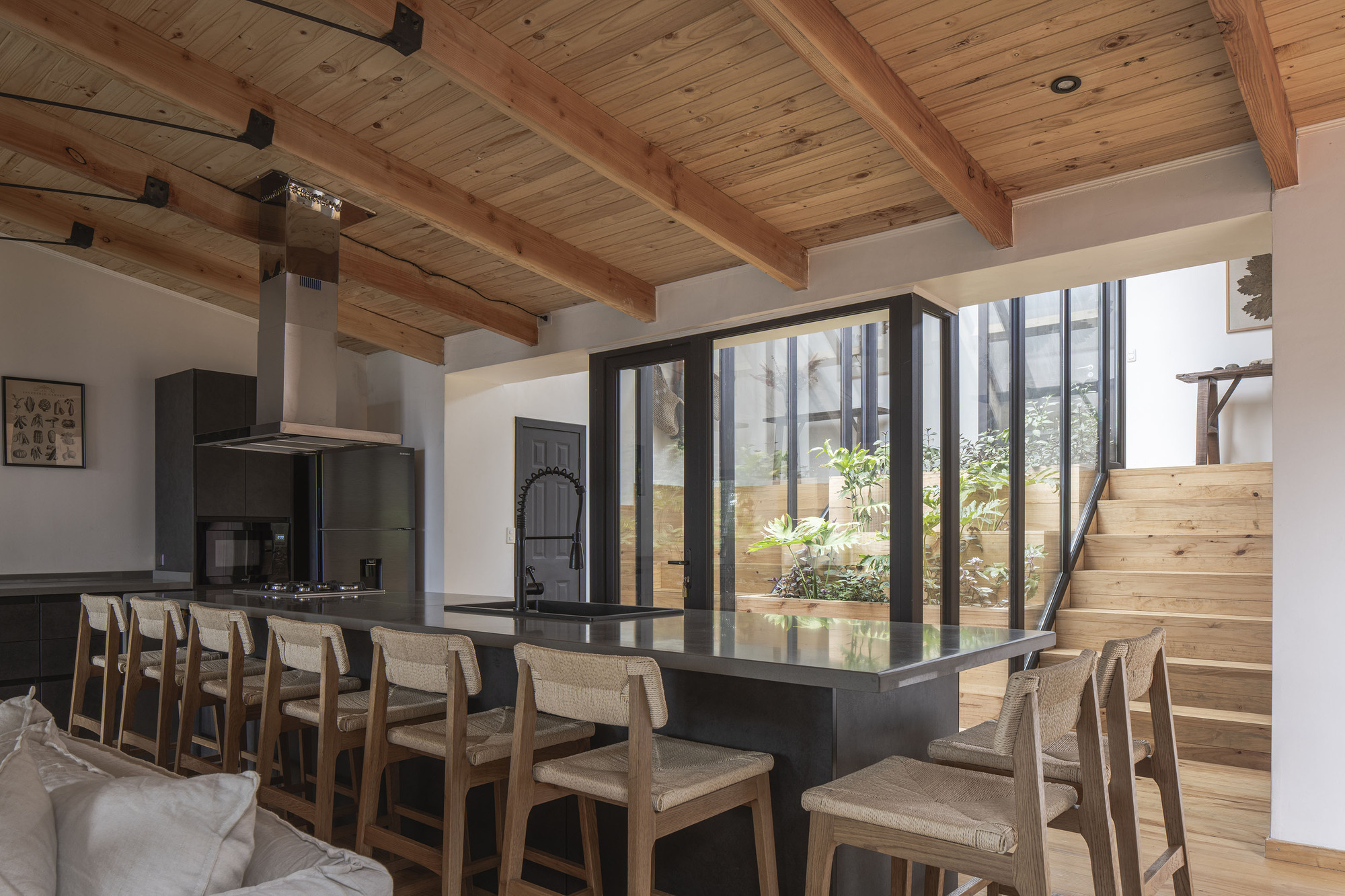 Dining area with black island and timber stairs beside floor-to-ceiling windows framing the trees outside