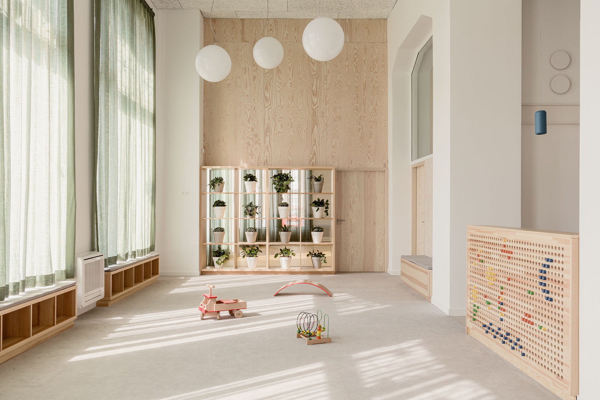 Double-height playroom with plywood plant wall, sphere pendant lights, and sheer curtains filtering daylight