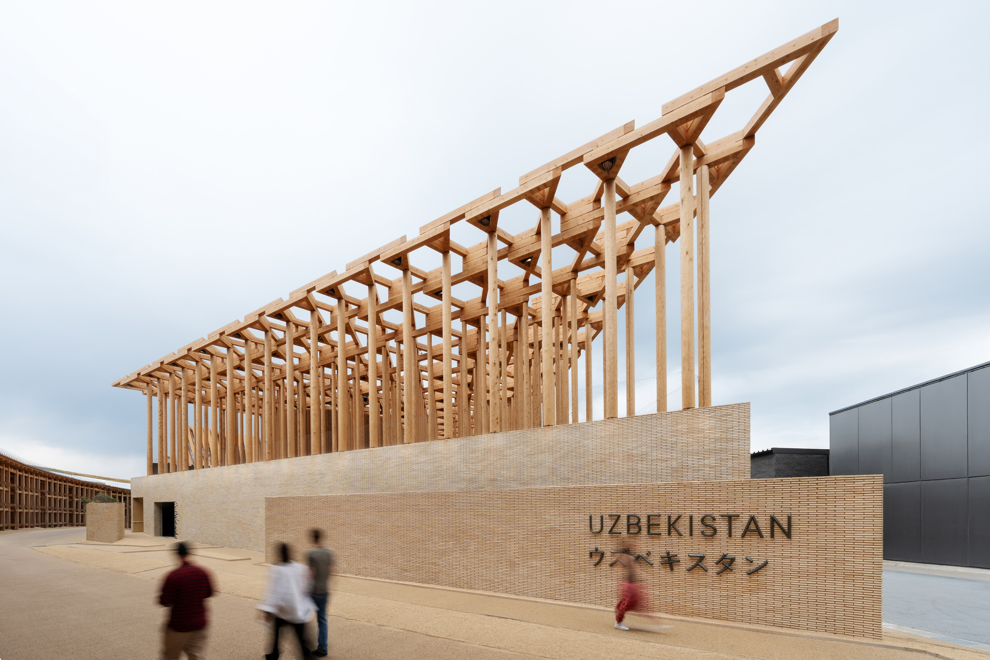 Diagonal timber roof structure rising above a beige brick facade with visitors walking past on an overcast day