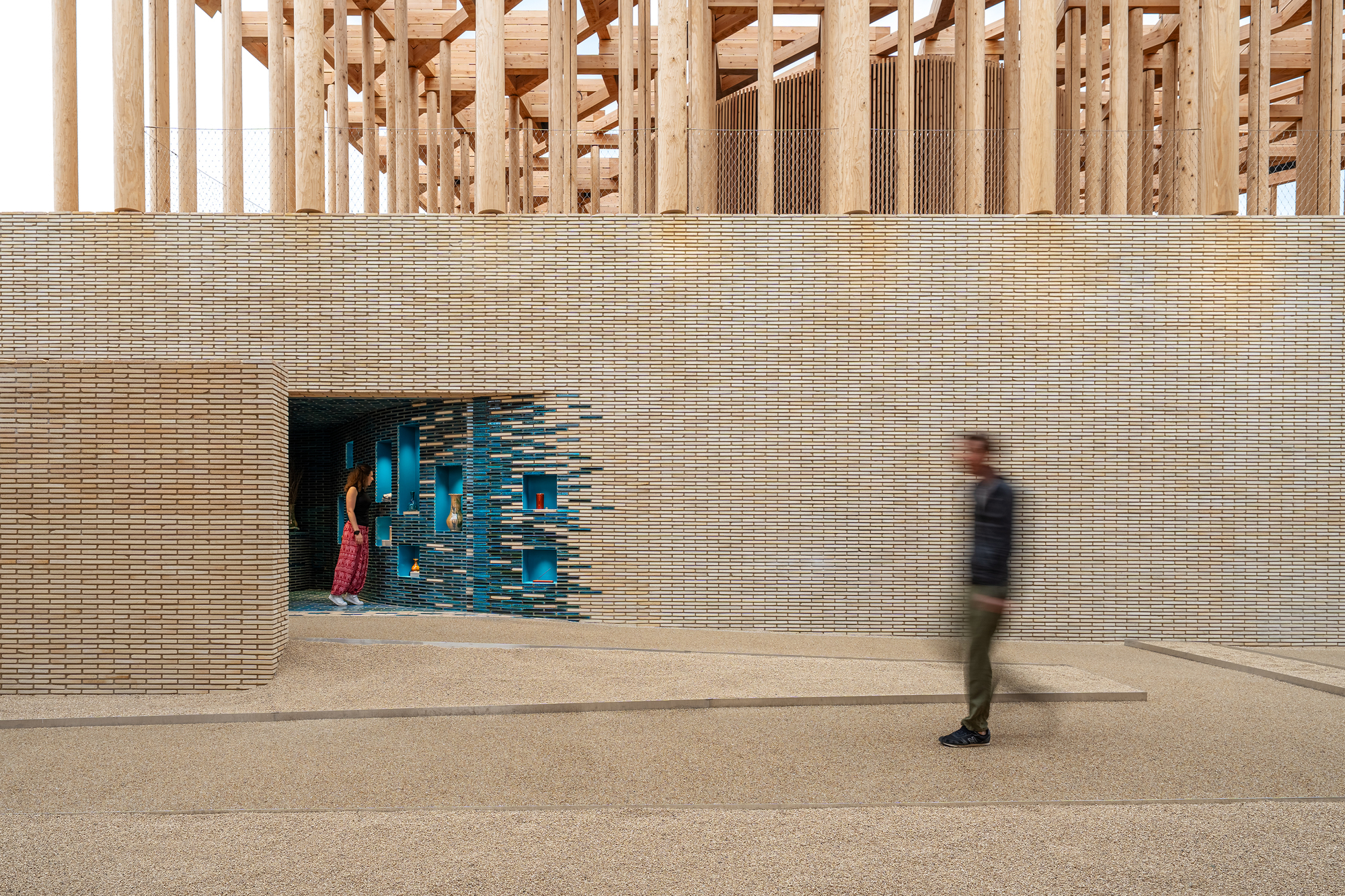 Beige brick wall with blue glazed tile installation around a doorway as a visitor walks past in afternoon light