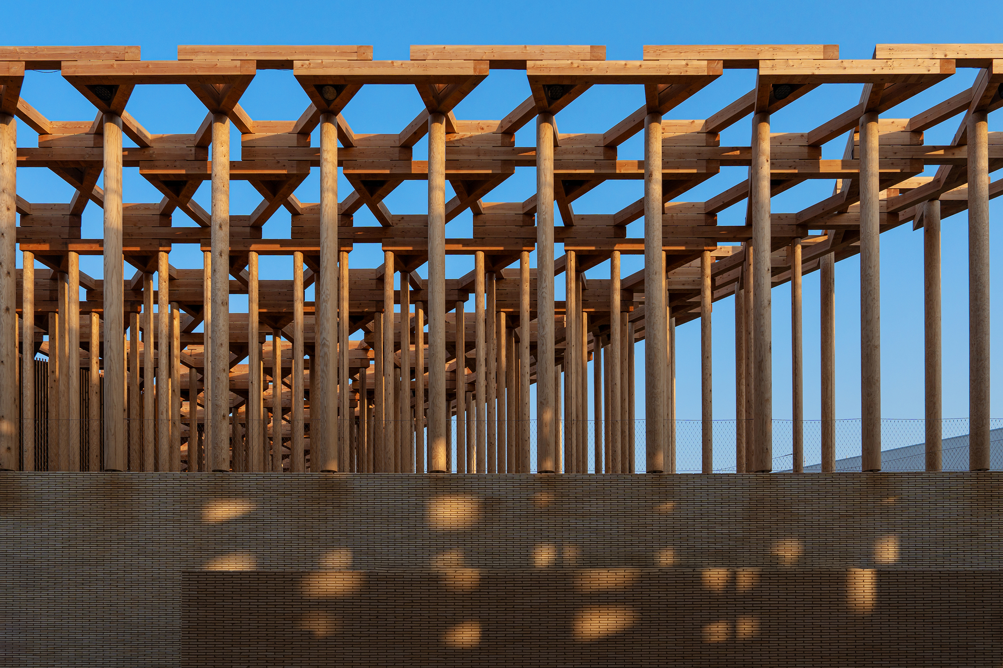Timber beam ceiling structure above beige brick wall casting geometric shadows in bright blue sky