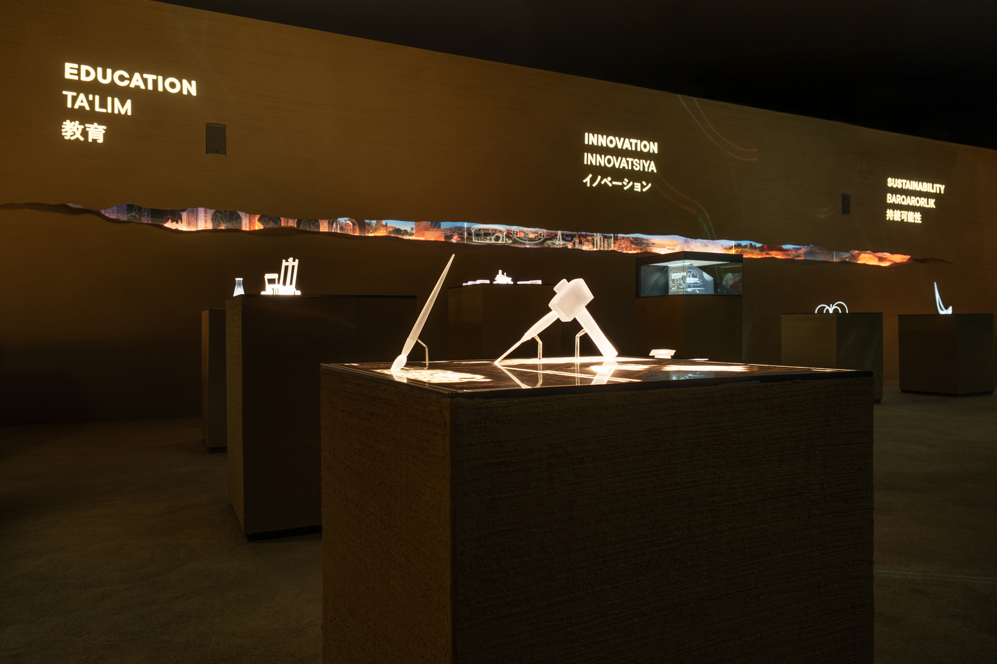 Exhibition tables with illuminated models beneath text panels in a darkened gallery space