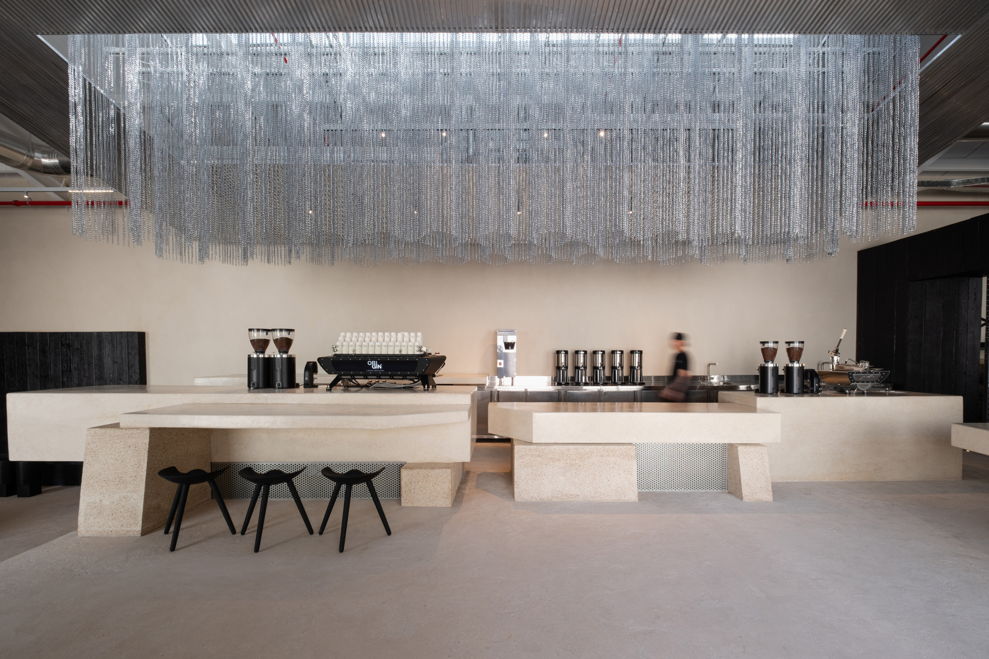 Long terrazzo service counter with espresso equipment beneath a cascading metal chain installation and a passing figure