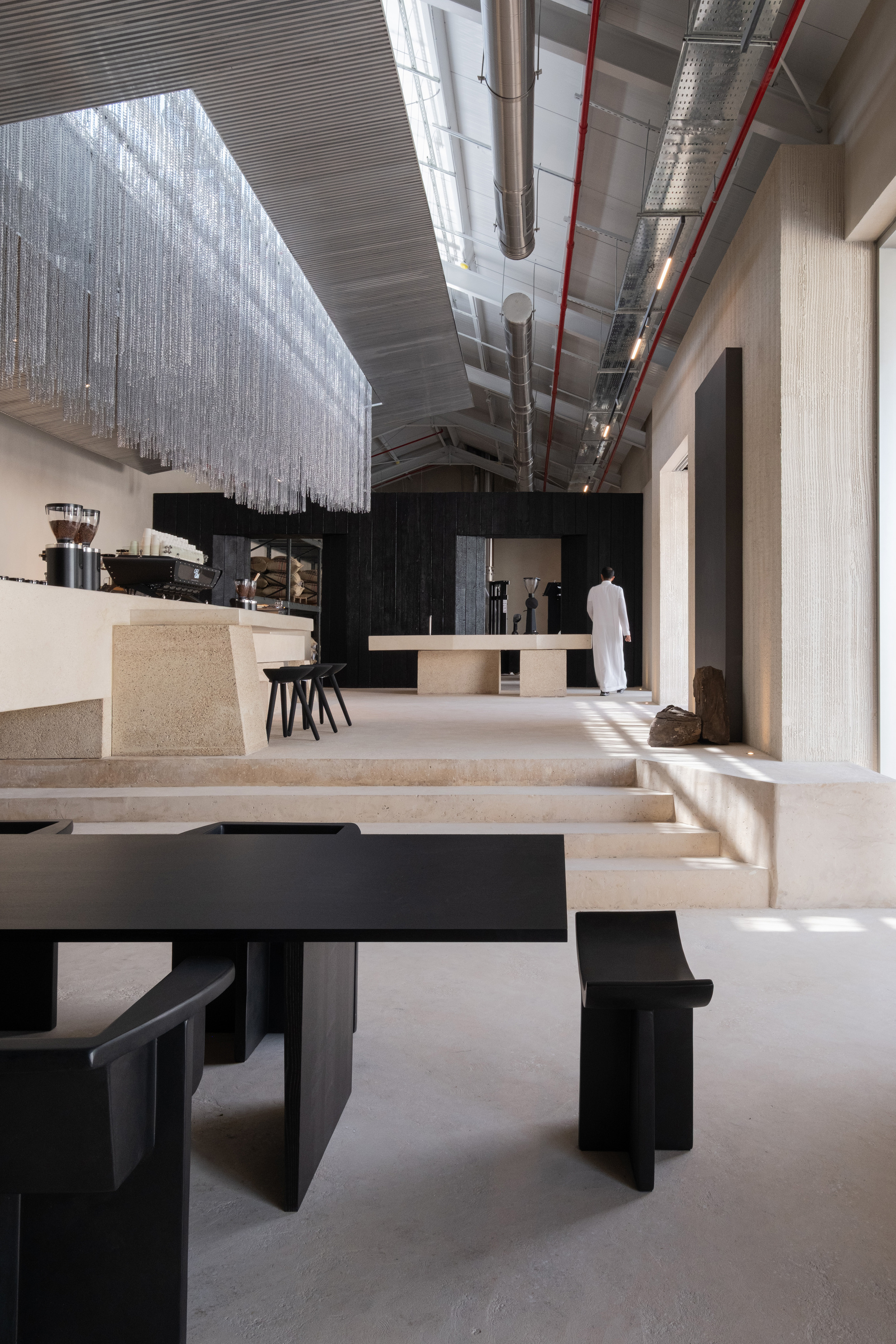 Interior view of tiered terrazzo seating with black tables beneath exposed ductwork and a translucent beaded curtain installation
