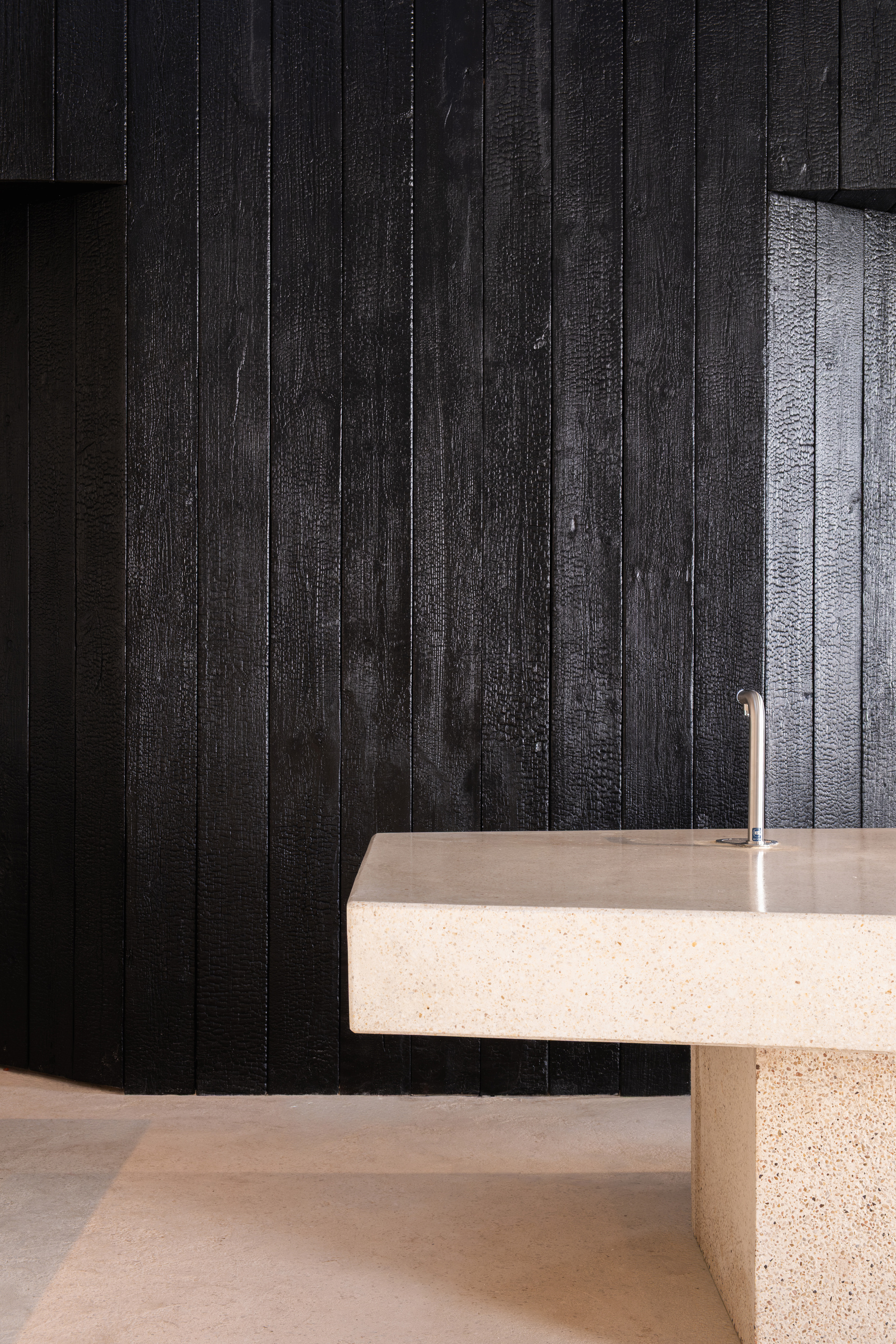 Terrazzo counter with cylindrical tap against charred timber cladding wall in darkened interior space