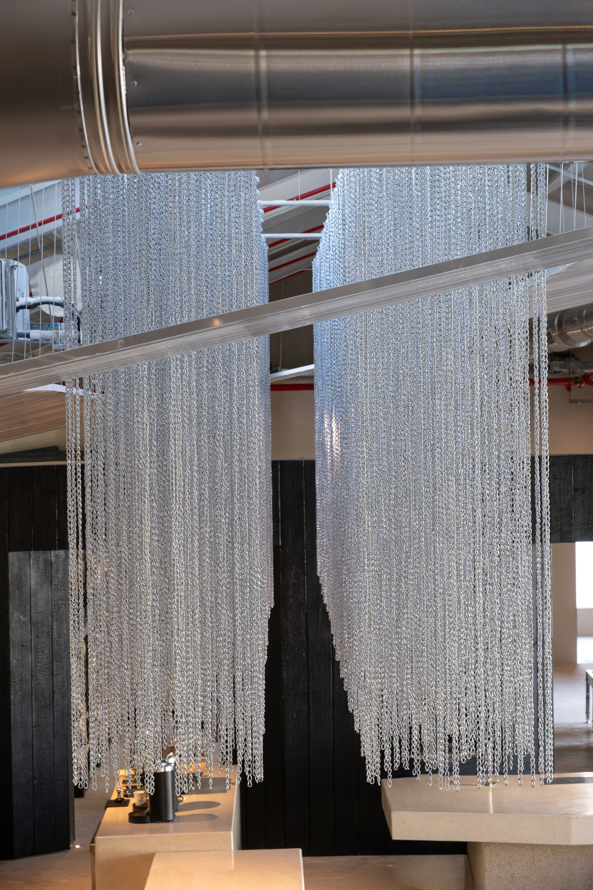 Suspended beaded chain curtain installation hanging from exposed ductwork and timber beams above black coffee bar volume