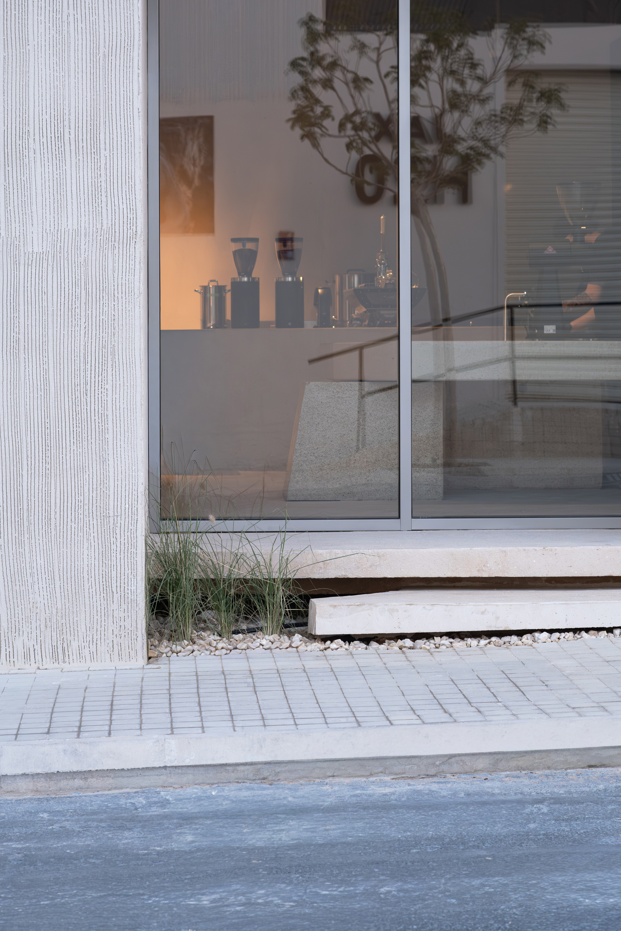 Glazed storefront threshold with textured concrete pilaster and pavers meeting gravel planting bed at dusk