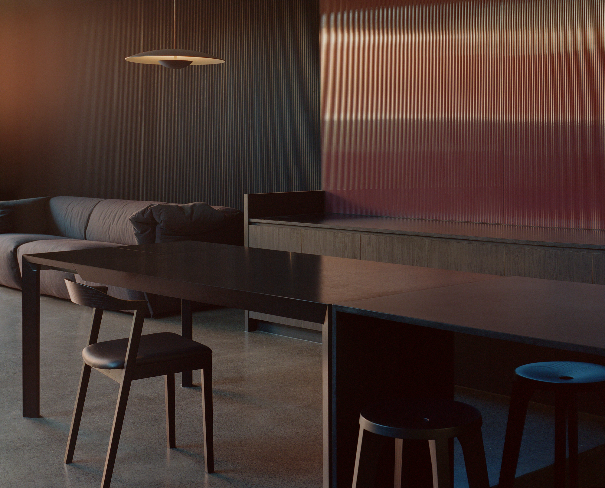 Dining table with timber chairs and stools at dusk with ribbed translucent wall glowing in background