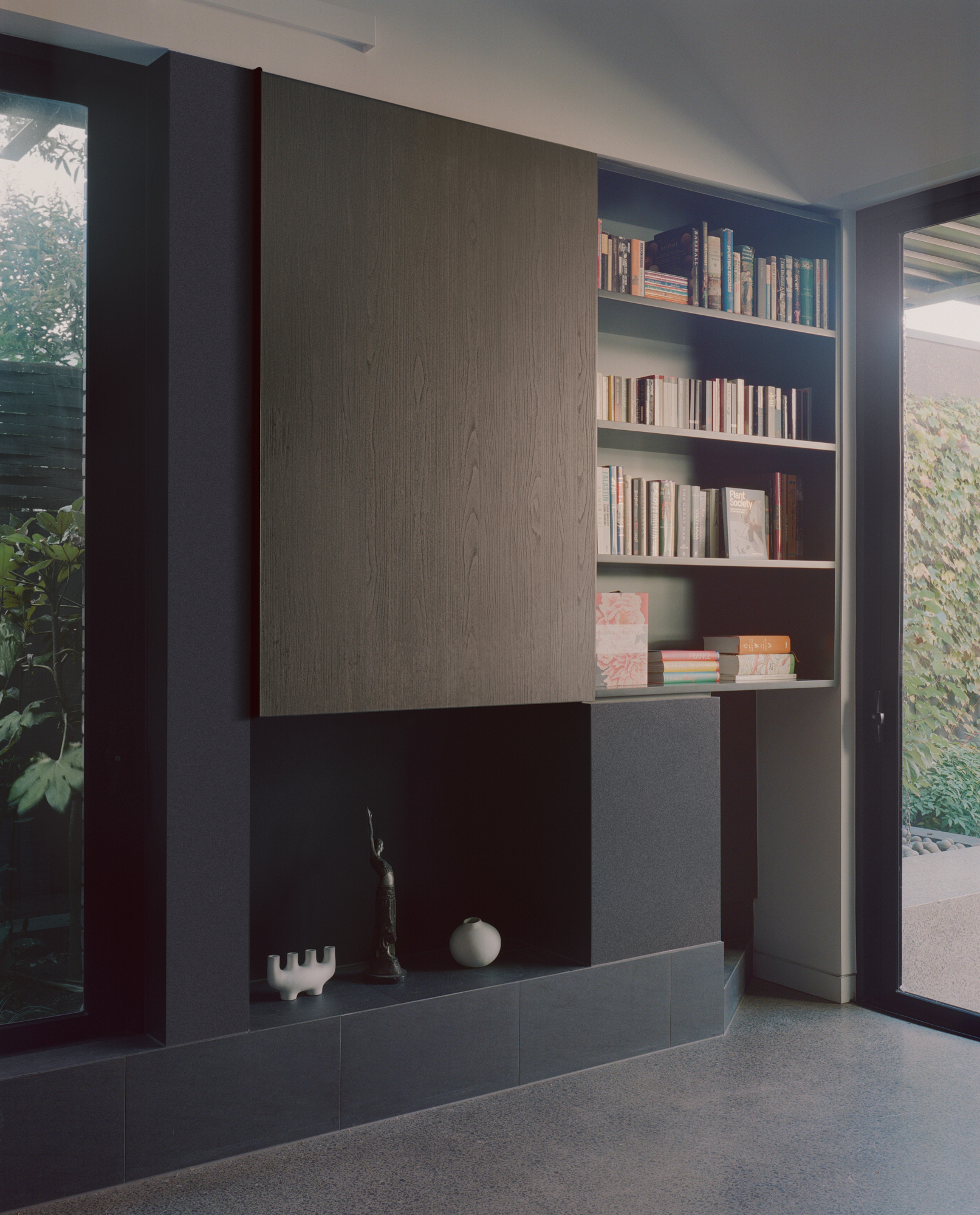Built-in bookshelf beside fireplace hearth with view to courtyard garden through glazed opening