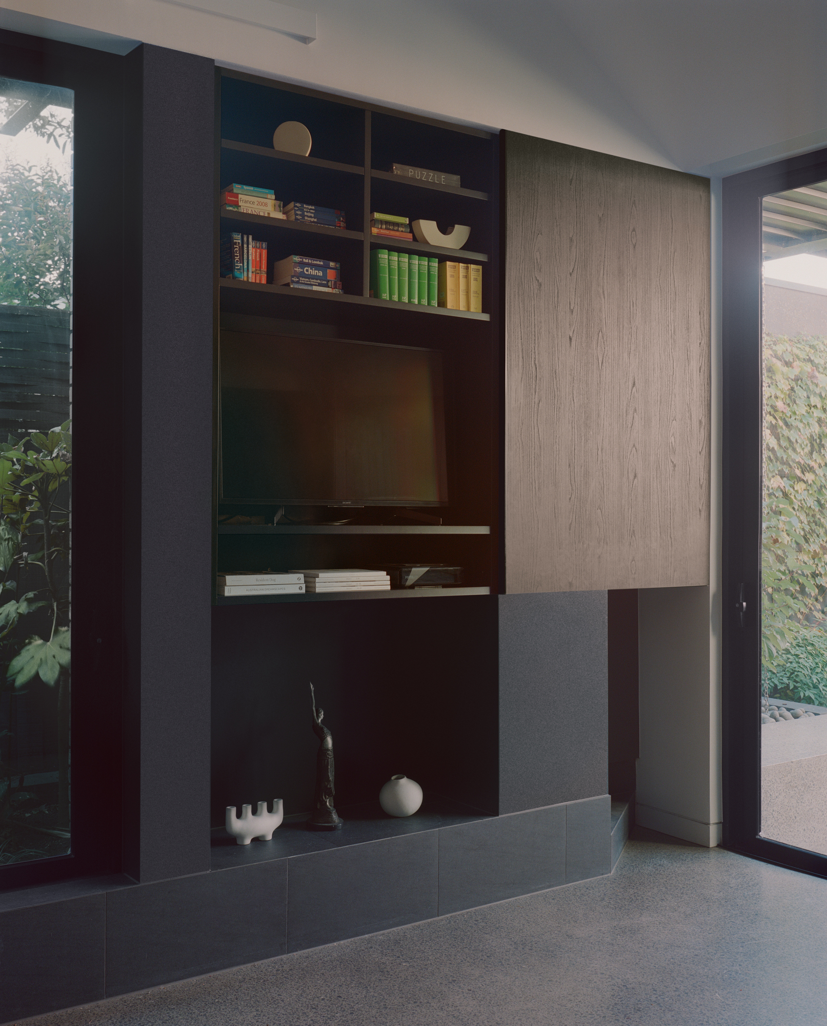 Built-in media unit with dark shelving and light oak cabinetry beside floor-to-ceiling glazing facing a garden